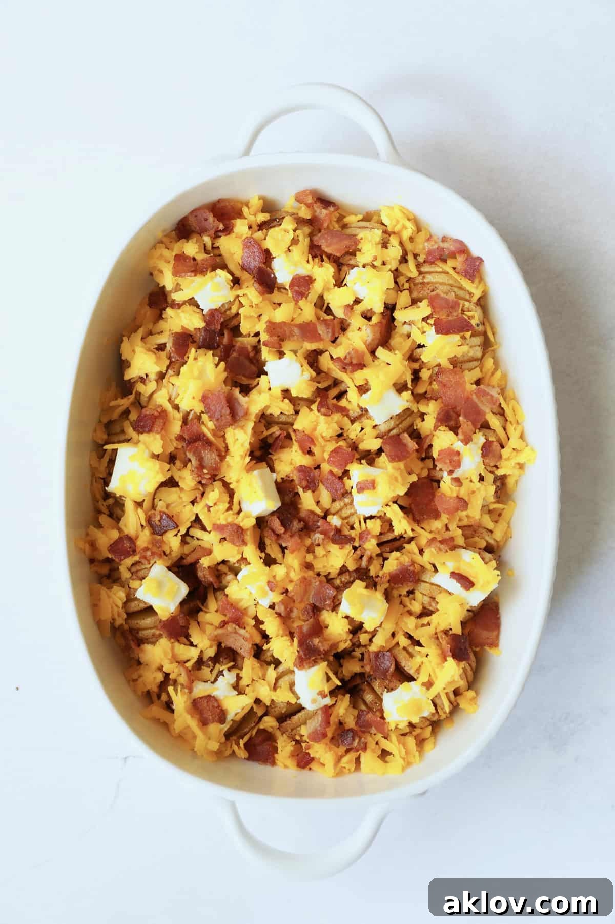 Photo of potatoes with butter, cheese and bacon in white oval baking dish ready to be baked.