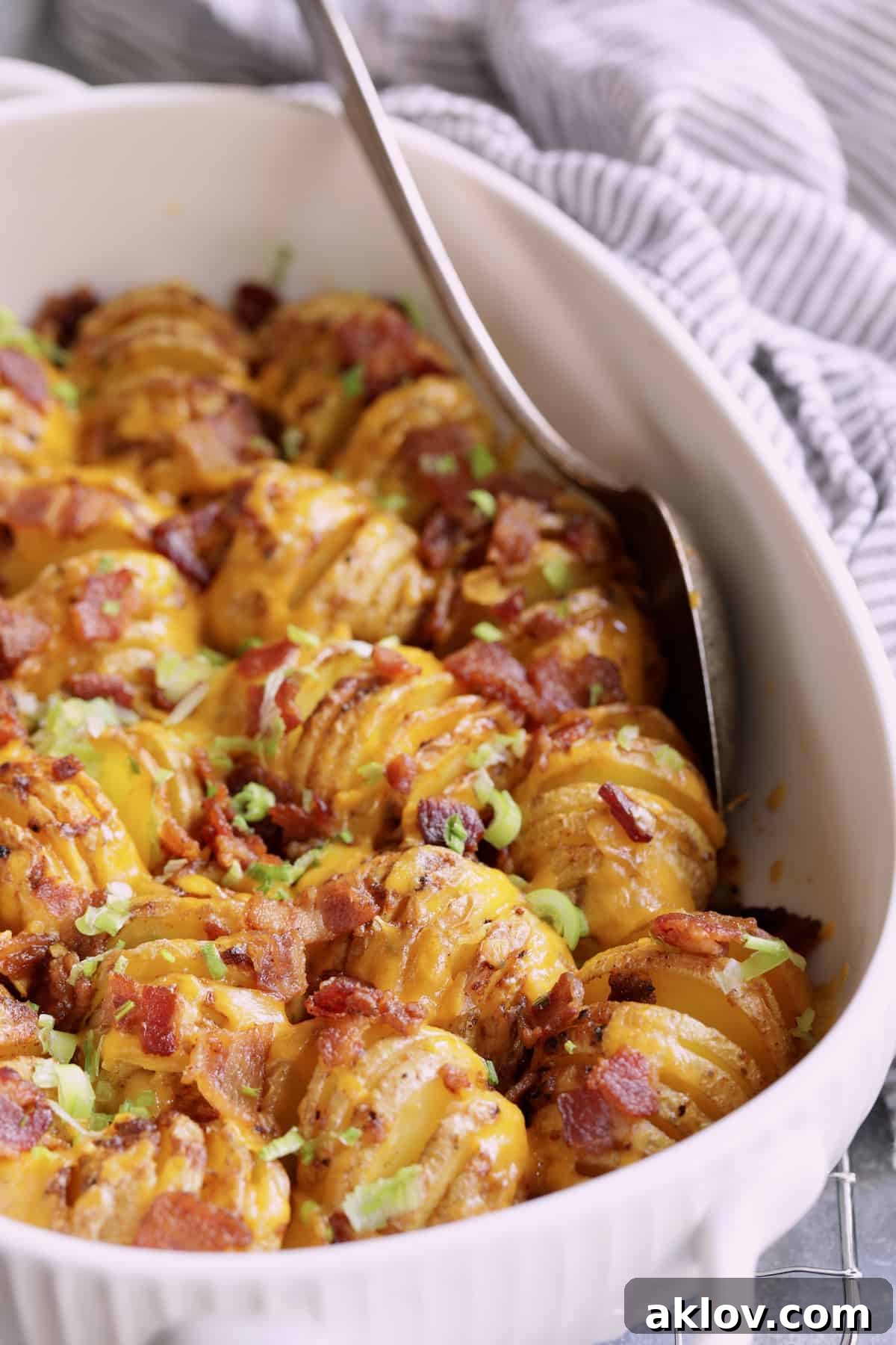 Close-up photo of Baby Hasselback Potato Bake with Cheddar and Bacon in white baking dish with serving spoon.