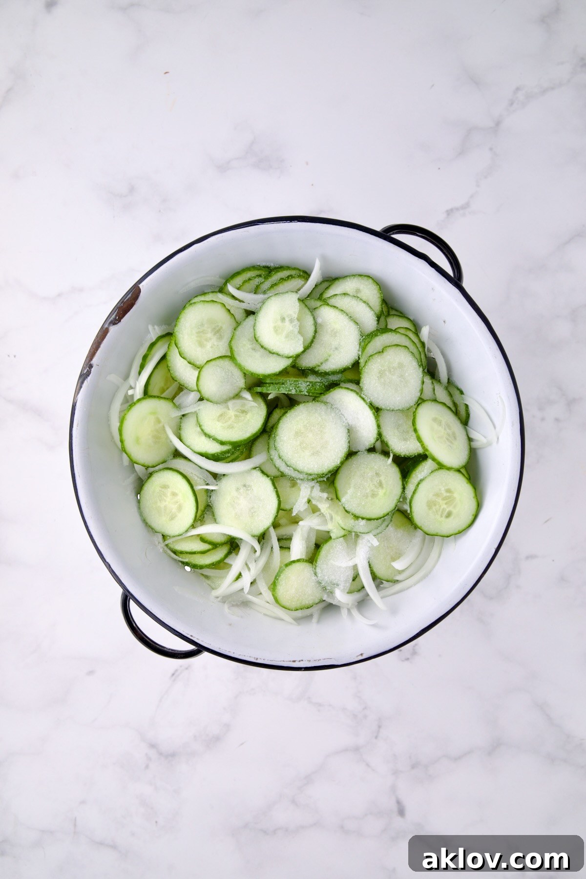 Cucumbers and onions in a white distressed colander sprinkled with salt, beginning the sweating process.