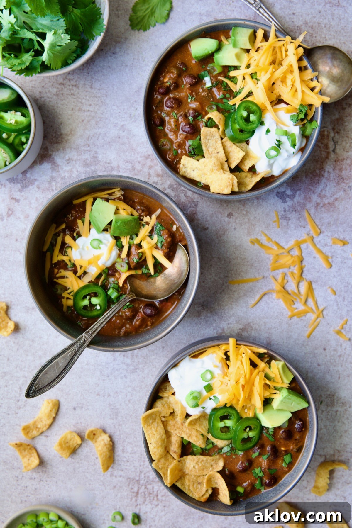 Three servings of Beef and Chorizo Chili in gray bowl garnished with avocado, cheddar cheese, sour cream, jalapeno slices and corn chips.