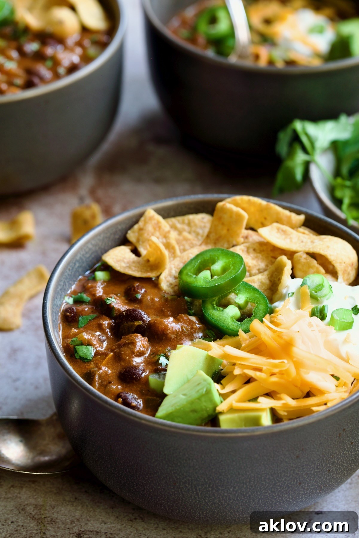 One serving of Beef and Chorizo Chili in gray bowl garnished with avocado, cheddar cheese, sour cream, jalapeno slices and corn chips.