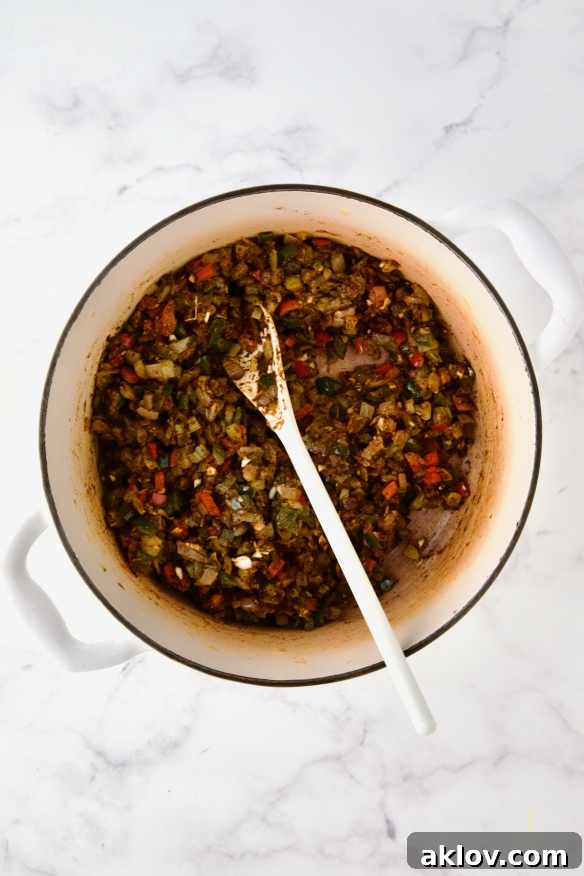 Spices stirred into cooked onions, celery, and peppers in white enamel Dutch oven with white mixing spoon.