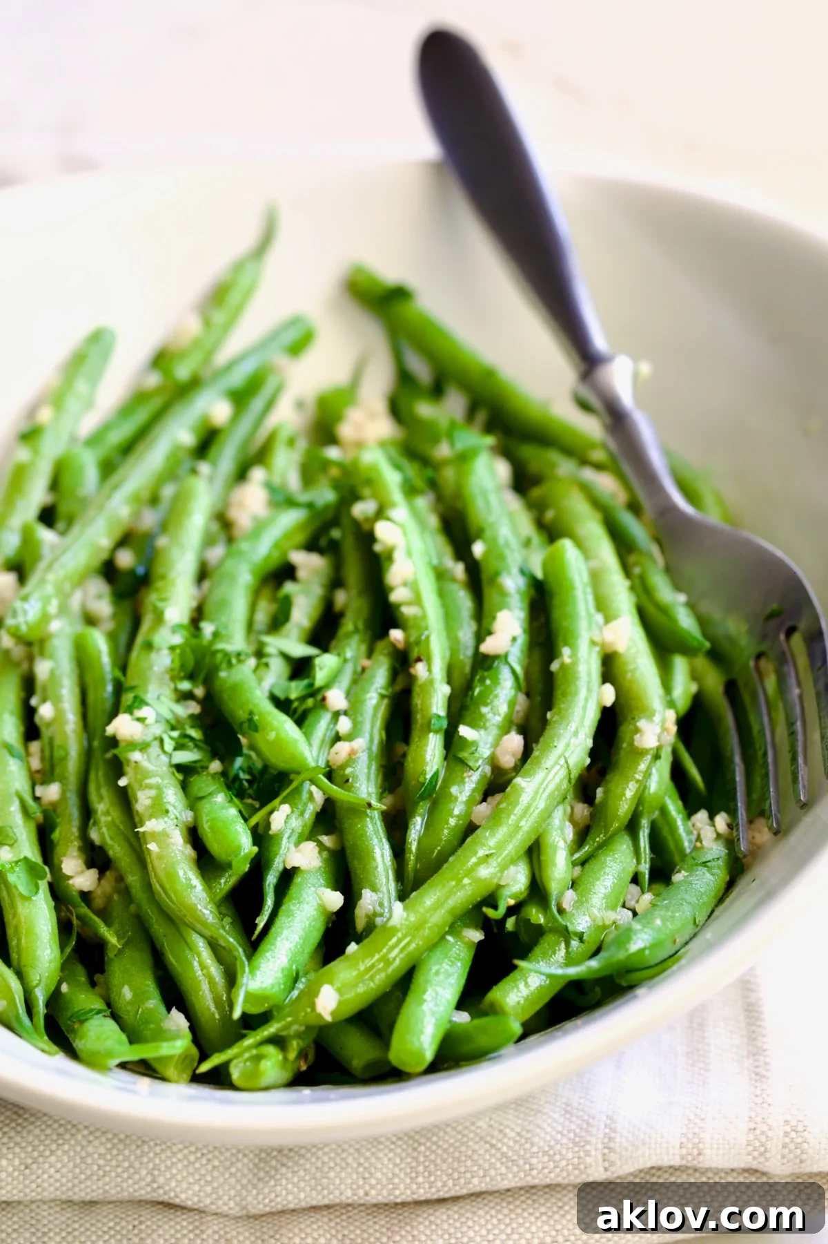 Garlic Green Beans in oval white serving bowl on folded beige towel with serving fork.