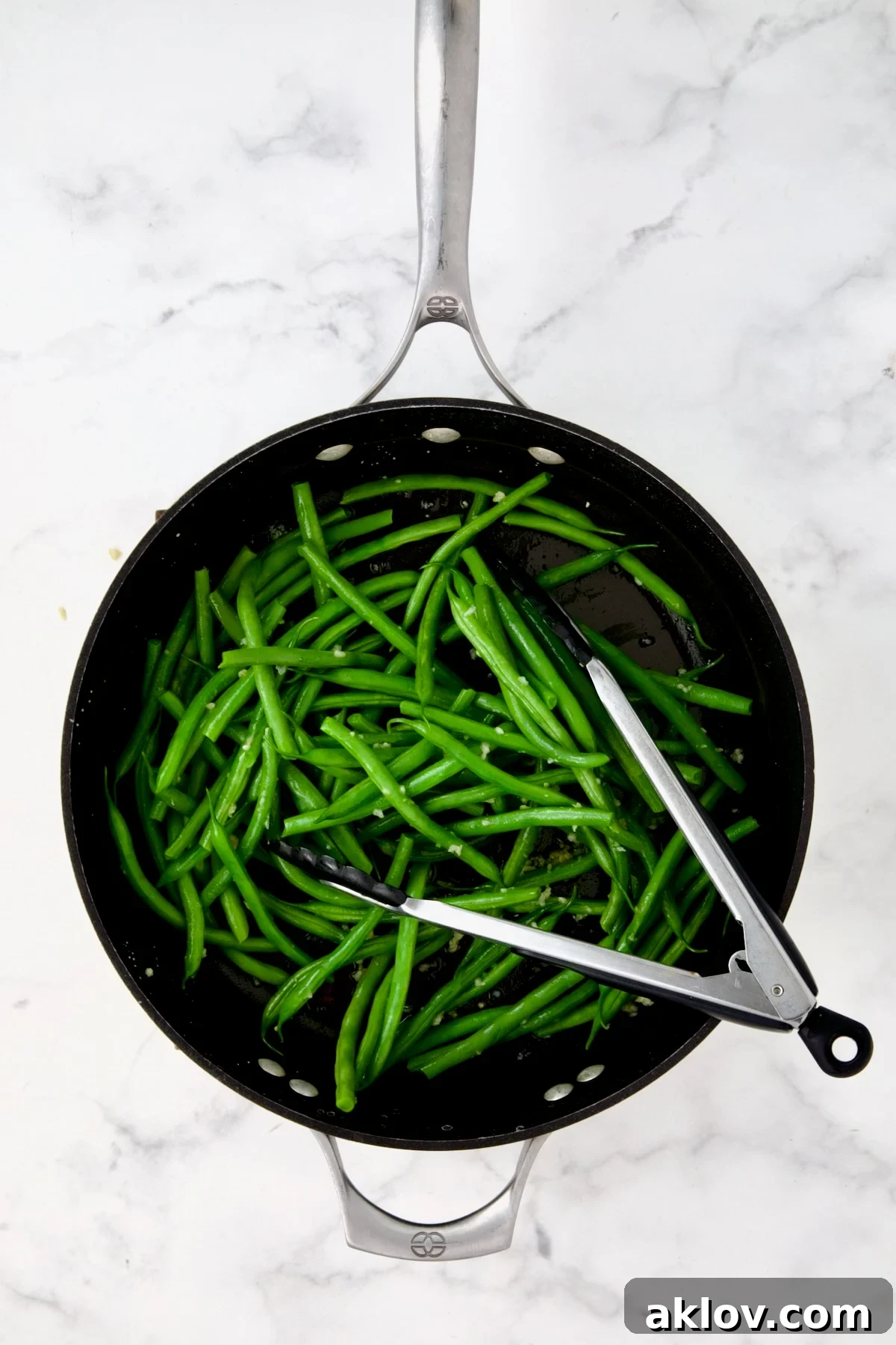 Green beans being tossed with sauteed garlic in nonstick skillet with stainless steel tongs.