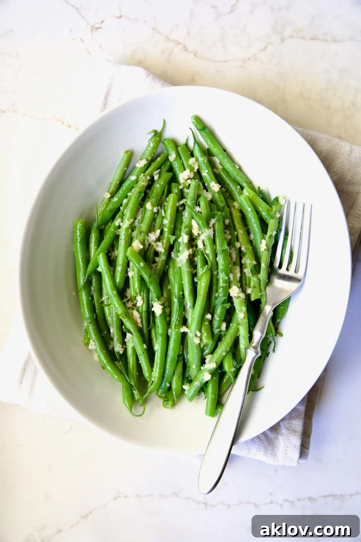 Garlic Green Beans in oval white serving bowl on folded beige towel with serving fork.