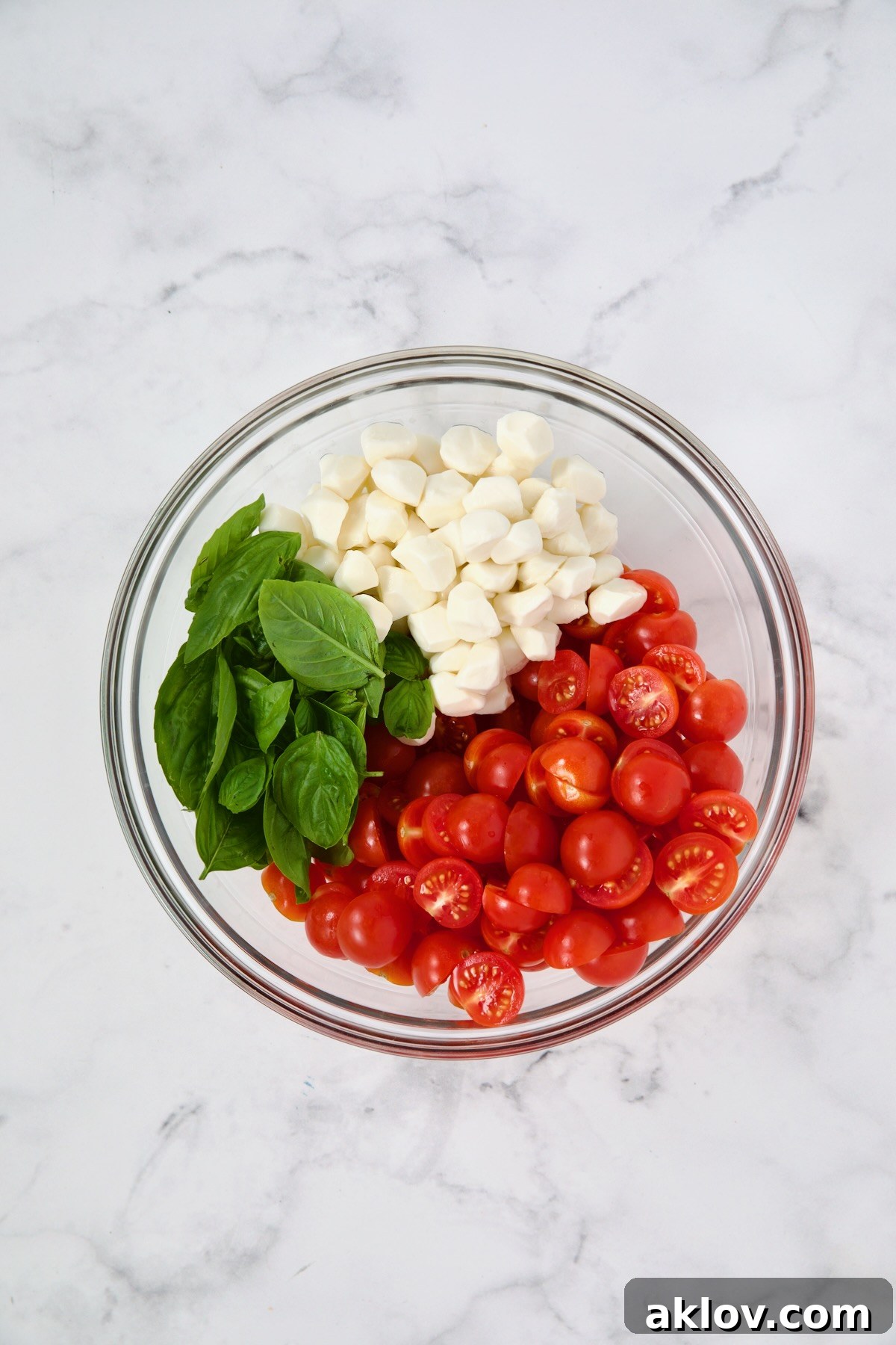 Fresh basil, mozzarella pearls, and halved cherry tomatoes neatly arranged in a glass bowl before being mixed.