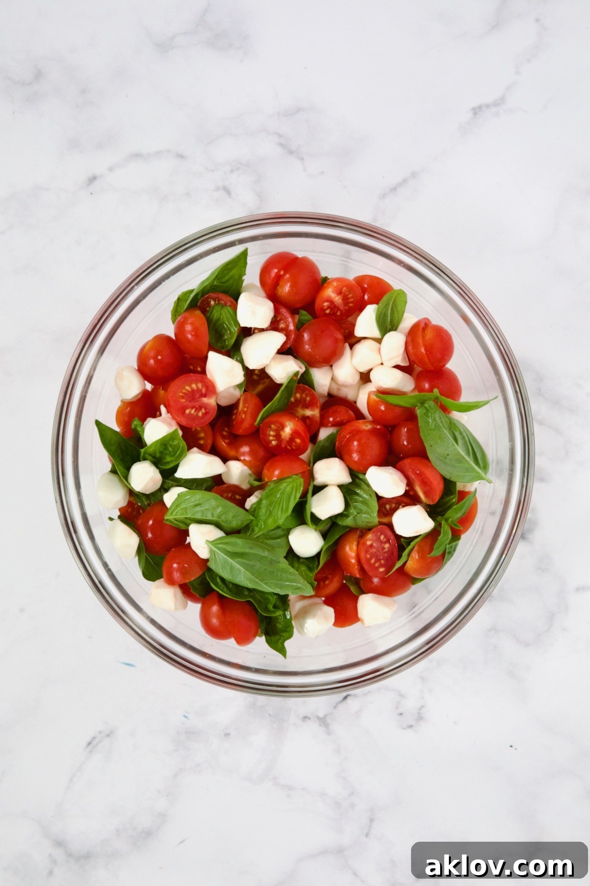 Fresh basil, mozzarella pearls, and halved cherry tomatoes gently mixed together in a glass bowl, ready for dressing.