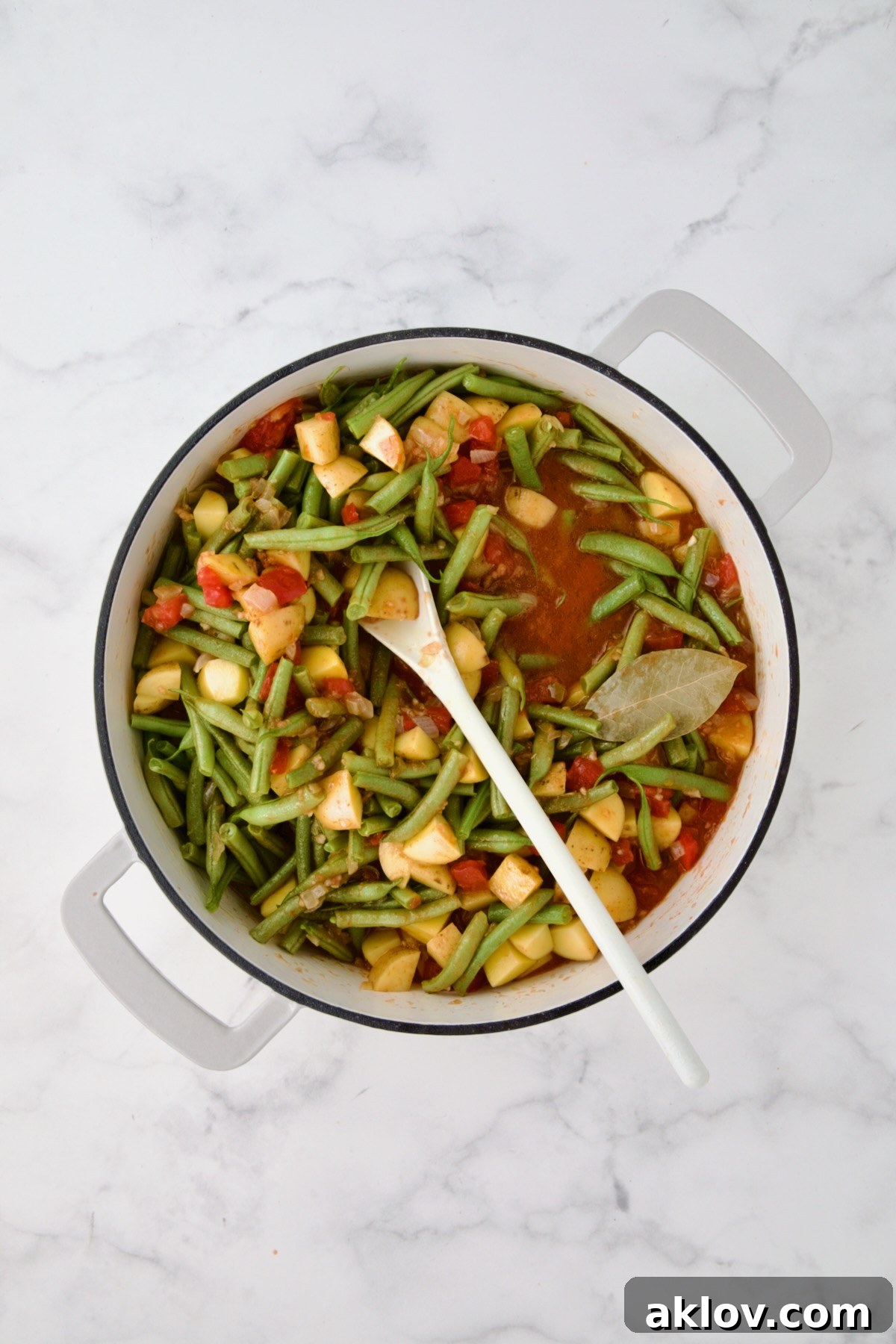 Potatoes, green beans and bay leaf added to tomato sauce being mixed together with white spoon in white enamel cast iron skillet.