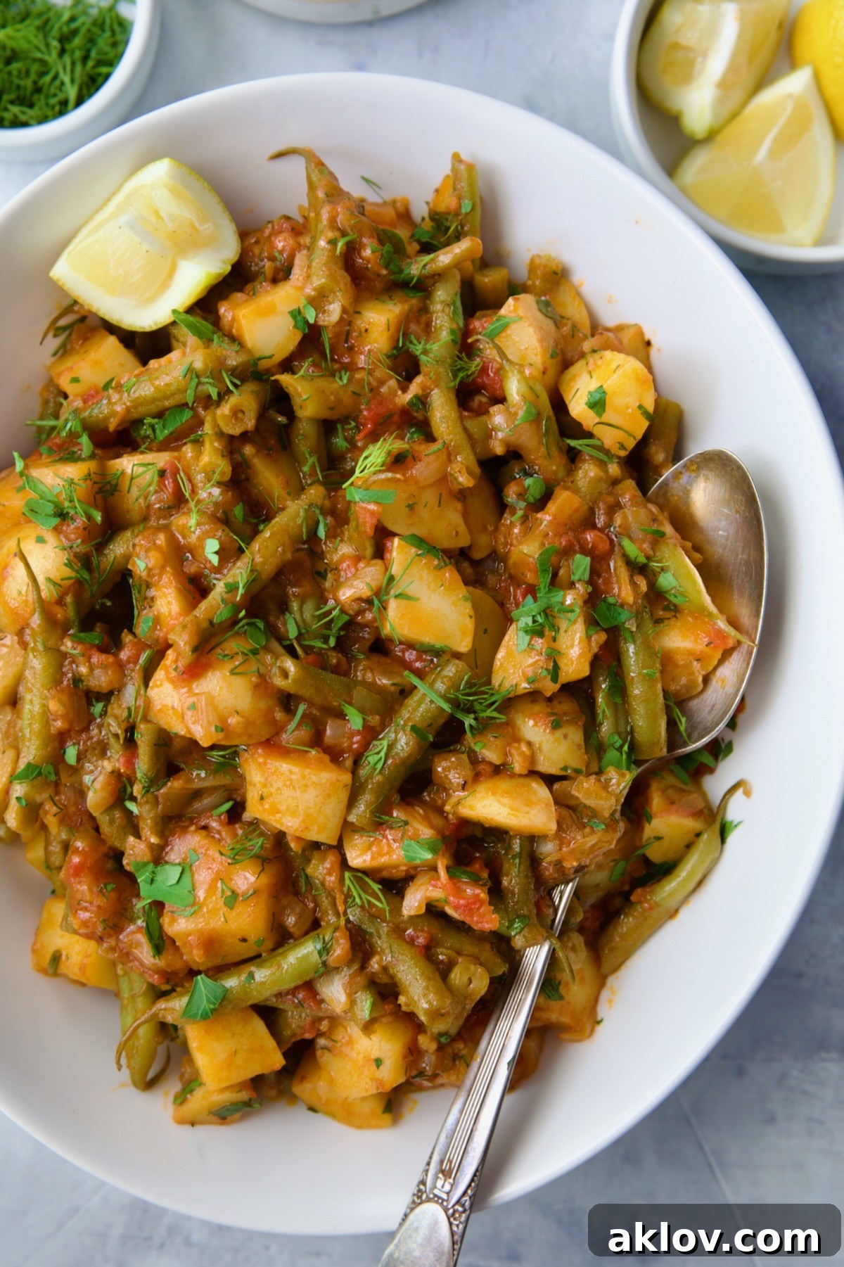 Fasolakia in oval white serving bowl garnished with fresh herbs and lemon wedges.