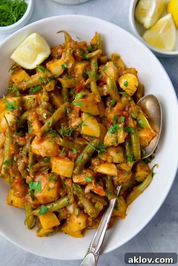 Fasolakia in oval white serving bowl garnished with fresh herbs and lemon wedges.