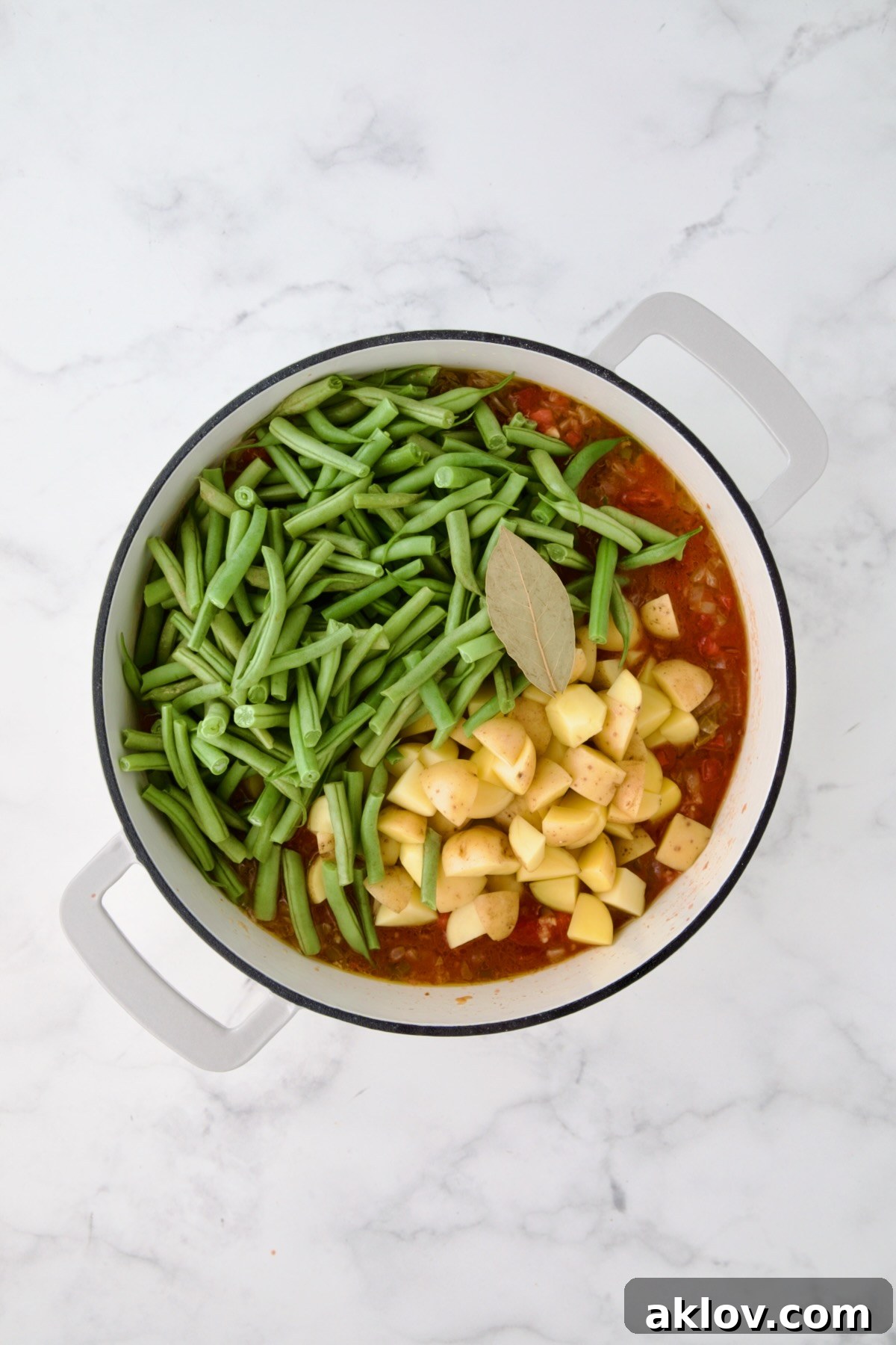 Potatoes, green beans and bay leaf added to tomato sauce in white enamel cast iron skillet.