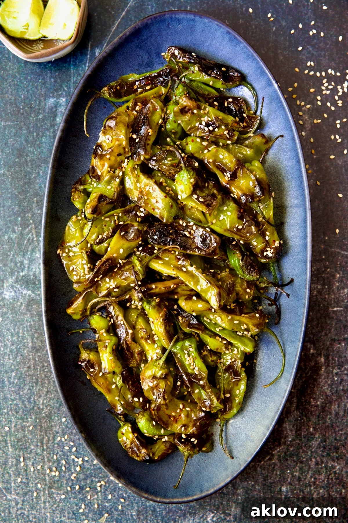 Blistered Shishito Peppers on a blue oval platter, garnished generously with toasted sesame seeds and lime wedges.