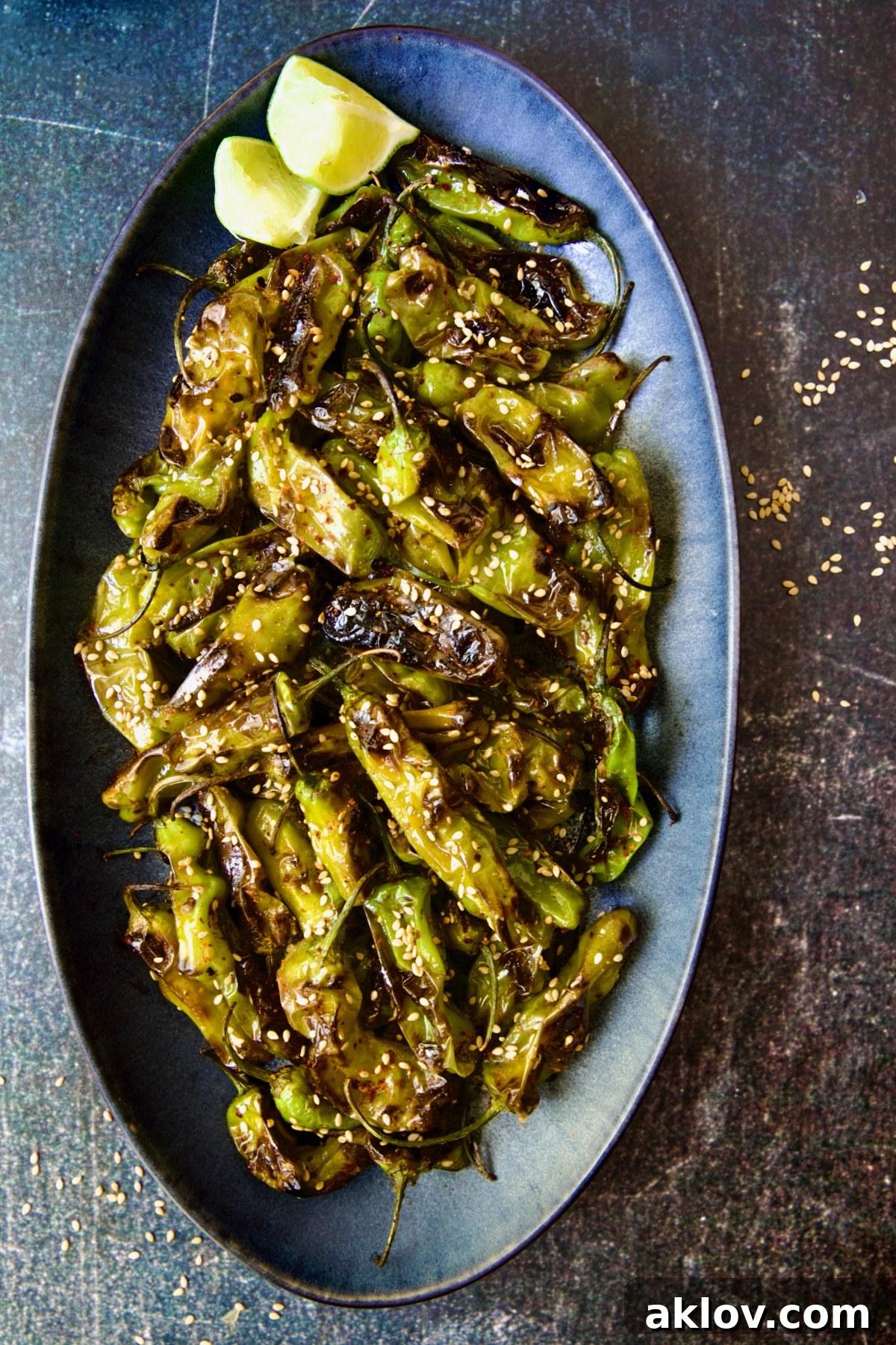 Blistered Shishito Peppers on a blue oval platter, garnished with sesame seeds and vibrant lime wedges for a fresh touch.