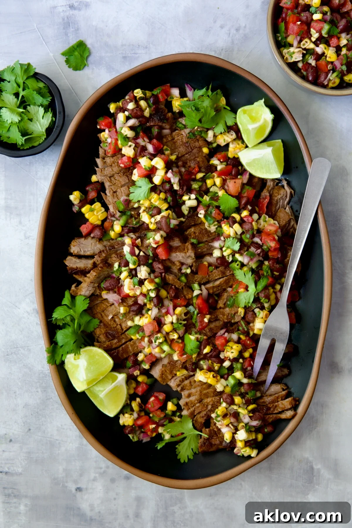 Grilled Skirt Steak with Black Bean Corn Tomato Salad on black platter garnished with lime wedges and cilantro sprigs.