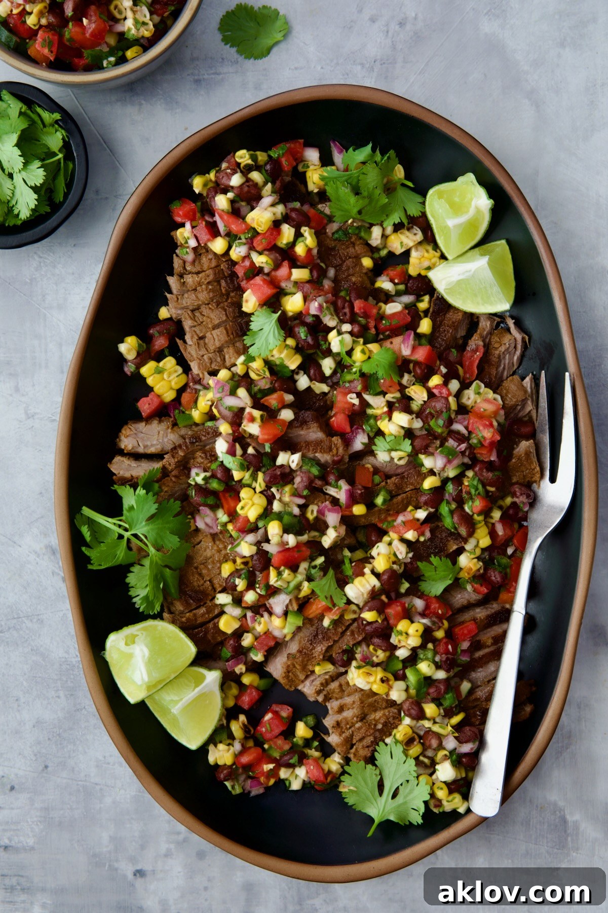 Grilled Skirt Steak with Black Bean Corn Tomato Salad on black platter garnished with lime wedges and cilantro sprigs.
