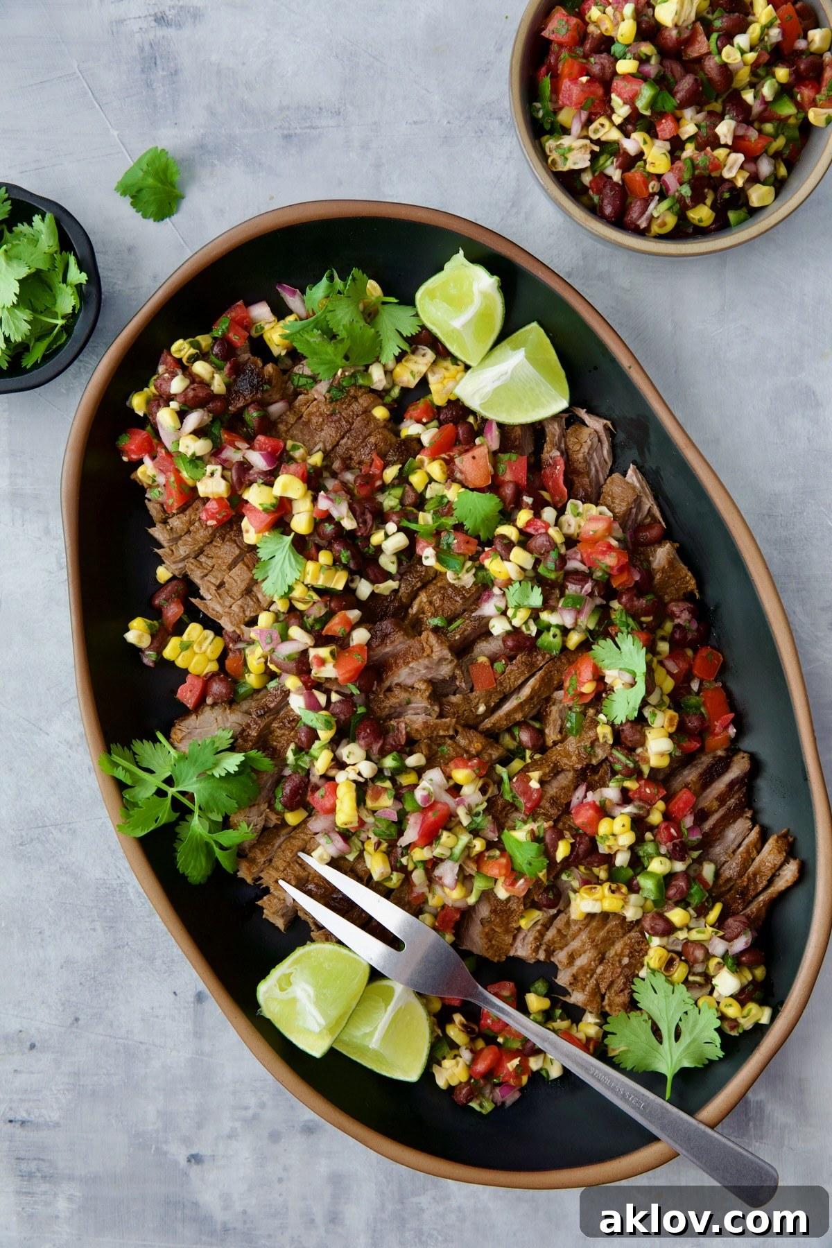 Grilled Skirt Steak with Black Bean Corn Tomato Salad on black platter garnished with lime wedges and cilantro sprigs.