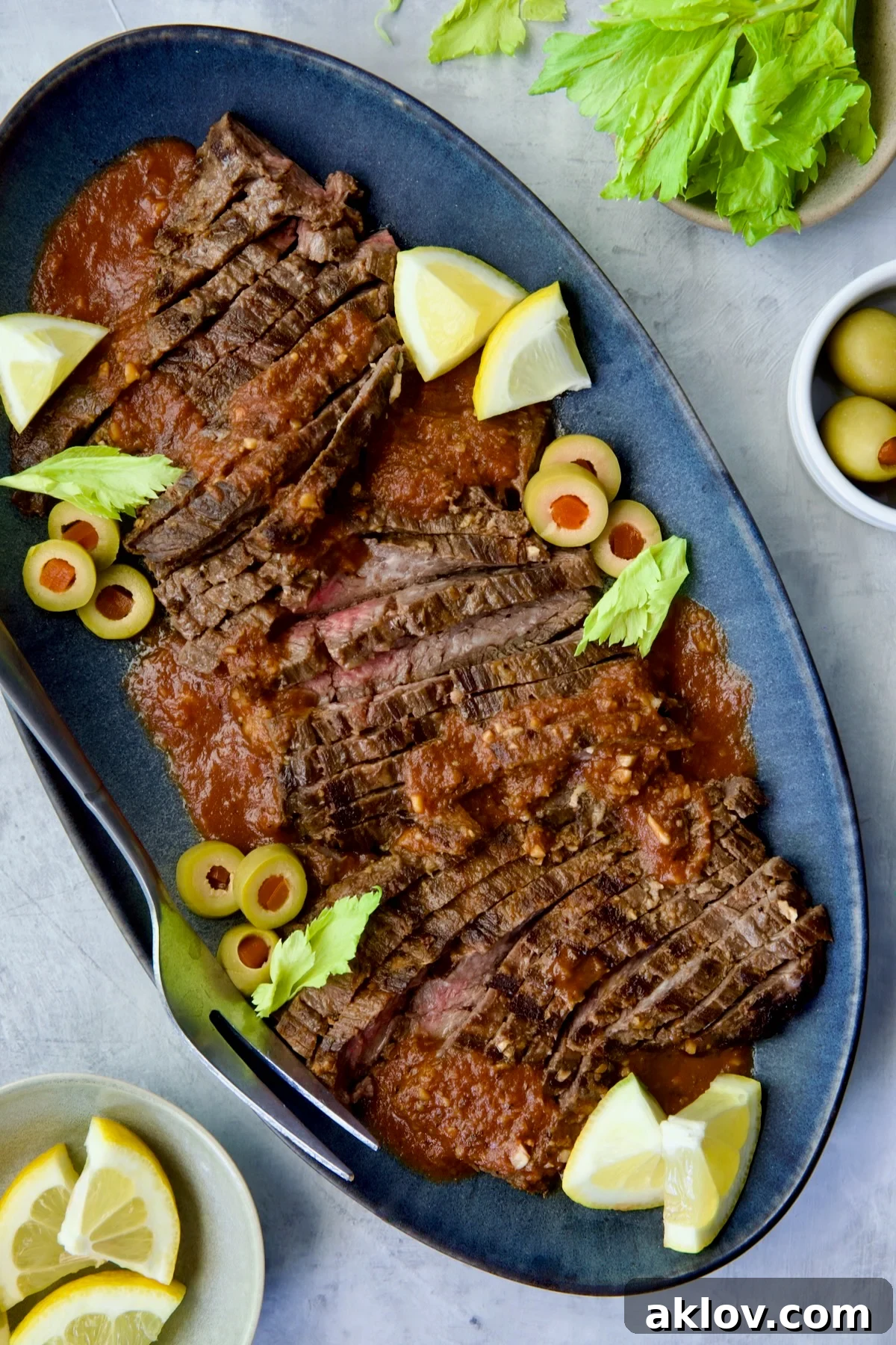 Sliced Bloody Mary Skirt Steak on blue oval platter garnished with lemon wedges, olive slices and celery leaves.