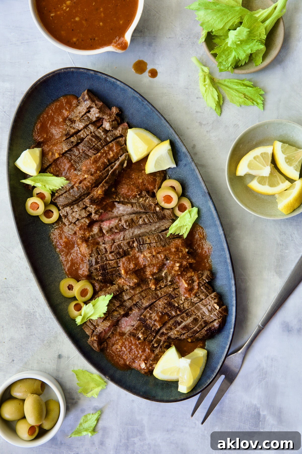 Sliced Bloody Mary Skirt Steak on blue oval platter garnished with lemon wedges, olive slices and celery leaves.