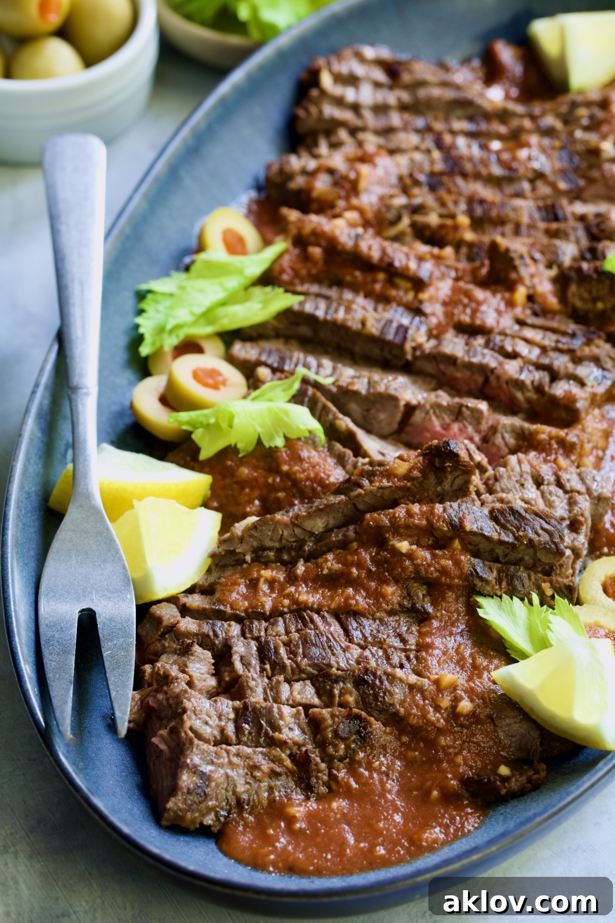 Sliced Bloody Mary Skirt Steak on blue oval platter garnished with lemon wedges, olive slices and celery leaves.