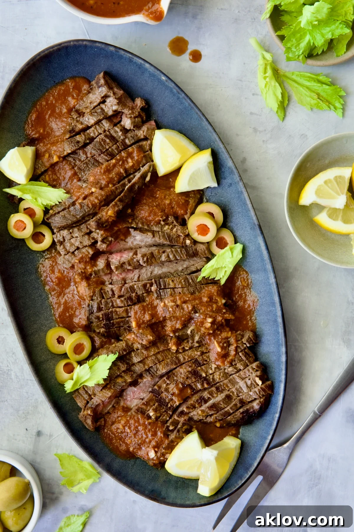 Sliced Bloody Mary Skirt Steak on blue oval platter garnished with lemon wedges, olive slices and celery leaves.