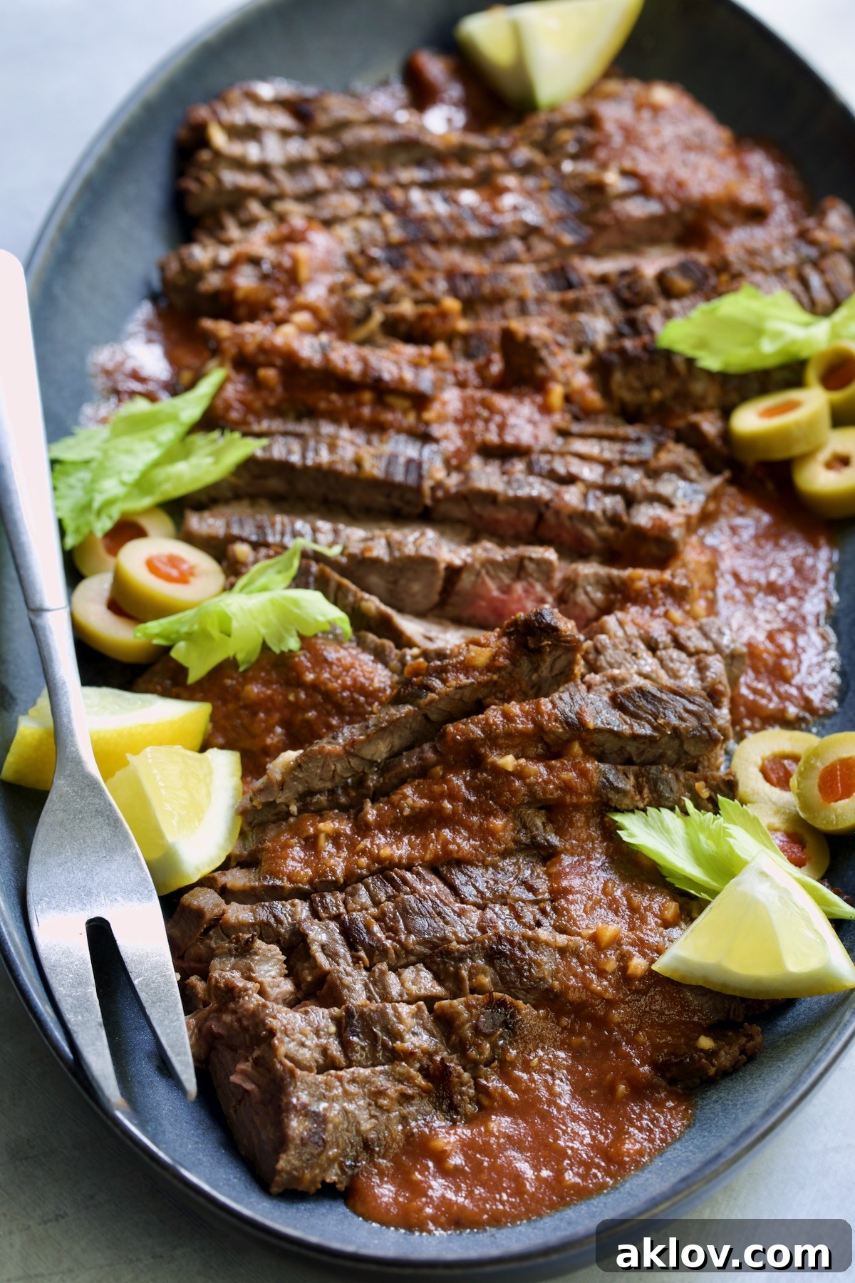 Sliced Bloody Mary Skirt Steak on blue oval platter garnished with lemon wedges, olive slices and celery leaves.