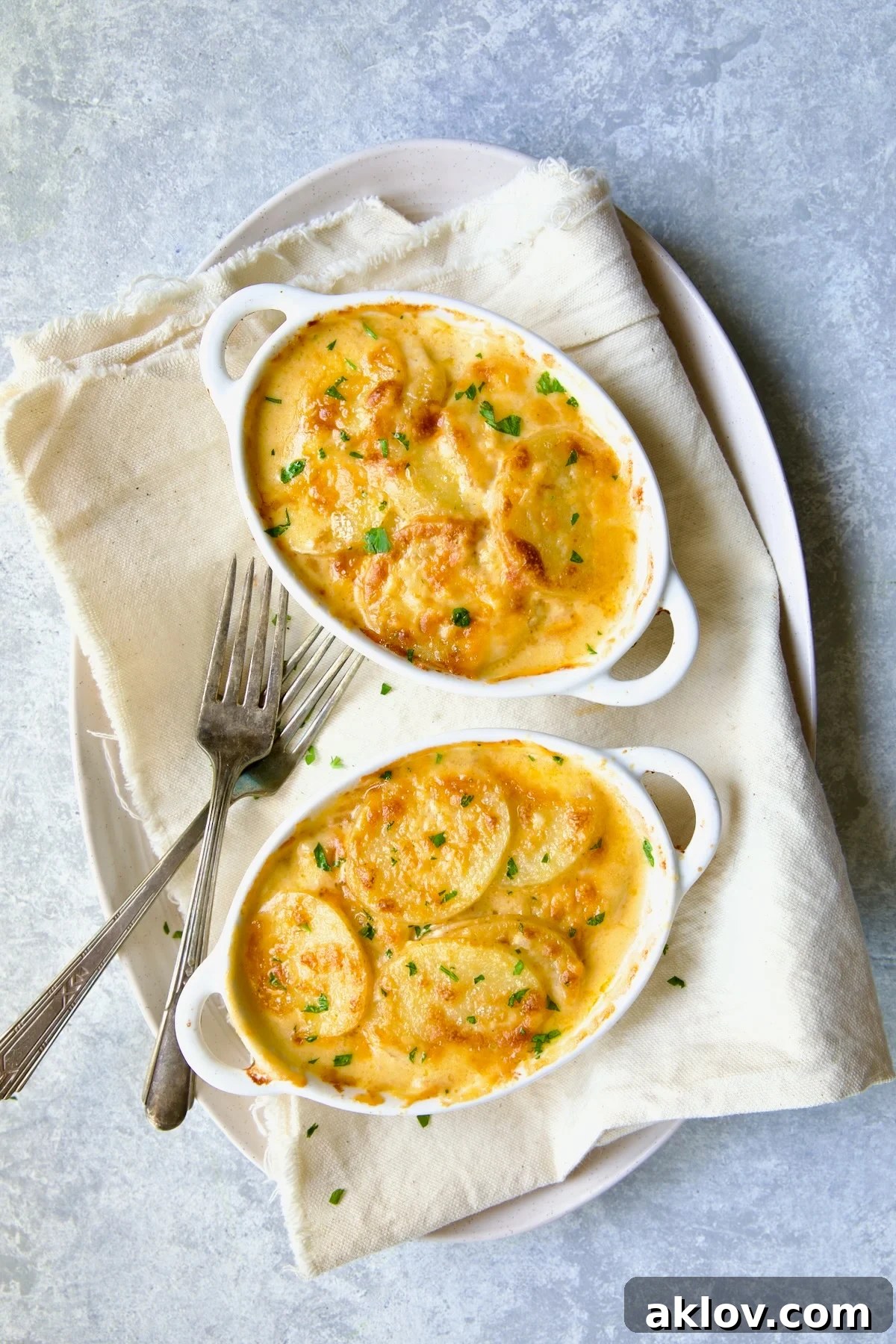 Two oval dishes of steaming hot Au Gratin Potatoes for Two, presented on a white oval platter with two forks, ready to serve.