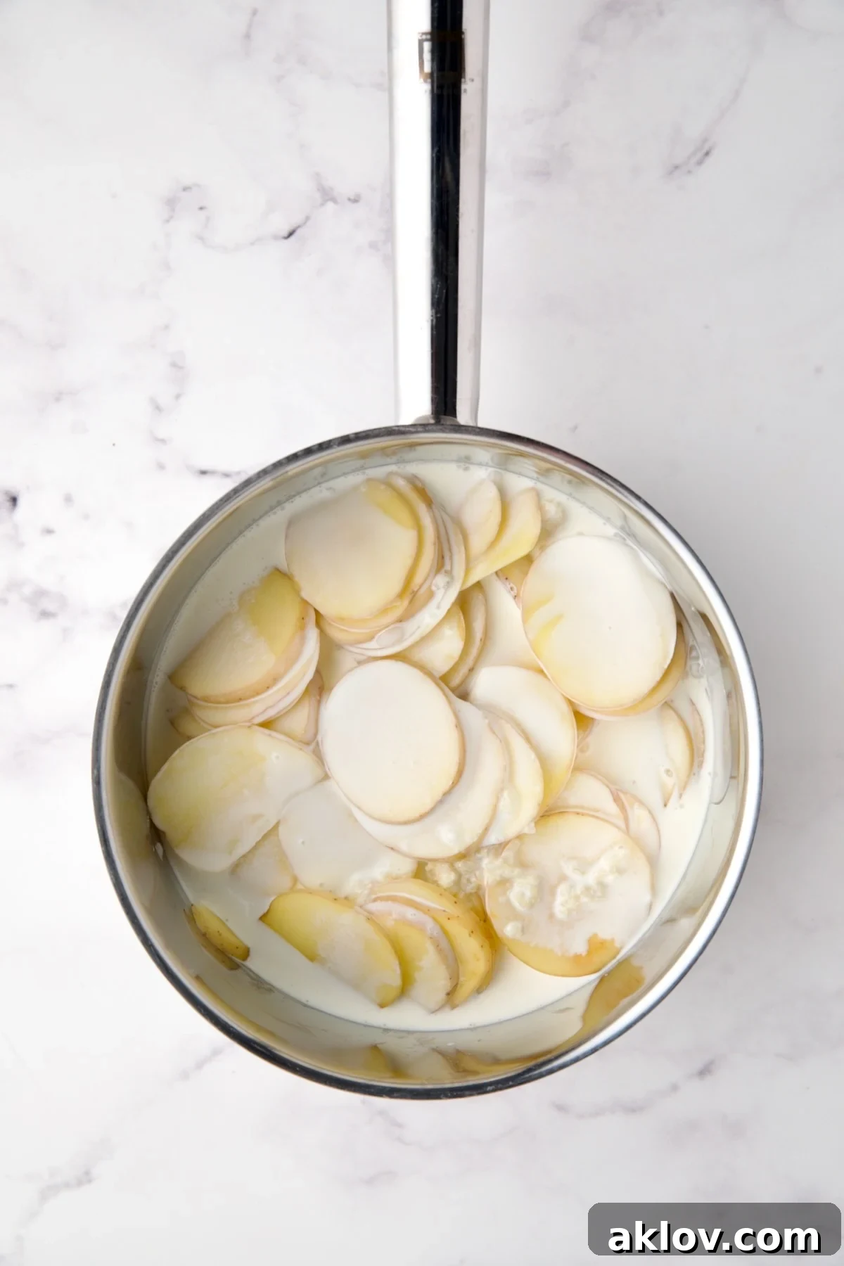 Rich heavy cream being poured over the potatoes, onion, and garlic in a stainless steel saucepan.