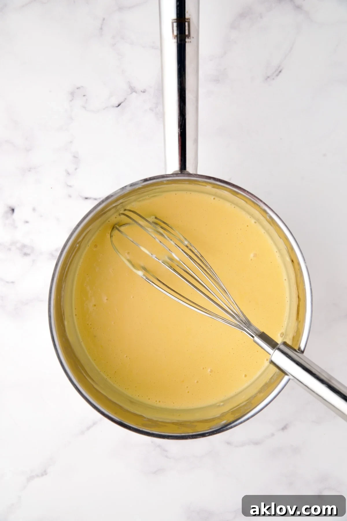 A wire whisk smoothly stirring a creamy, golden cheese sauce in a stainless steel saucepan.