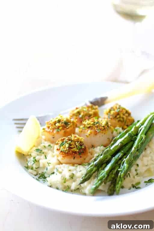 Pistachio Crusted Sea Scallops with Champagne Risotto and Roasted Asparagus on a white plate garnished with a lemon wedge and a wine glass in the background.