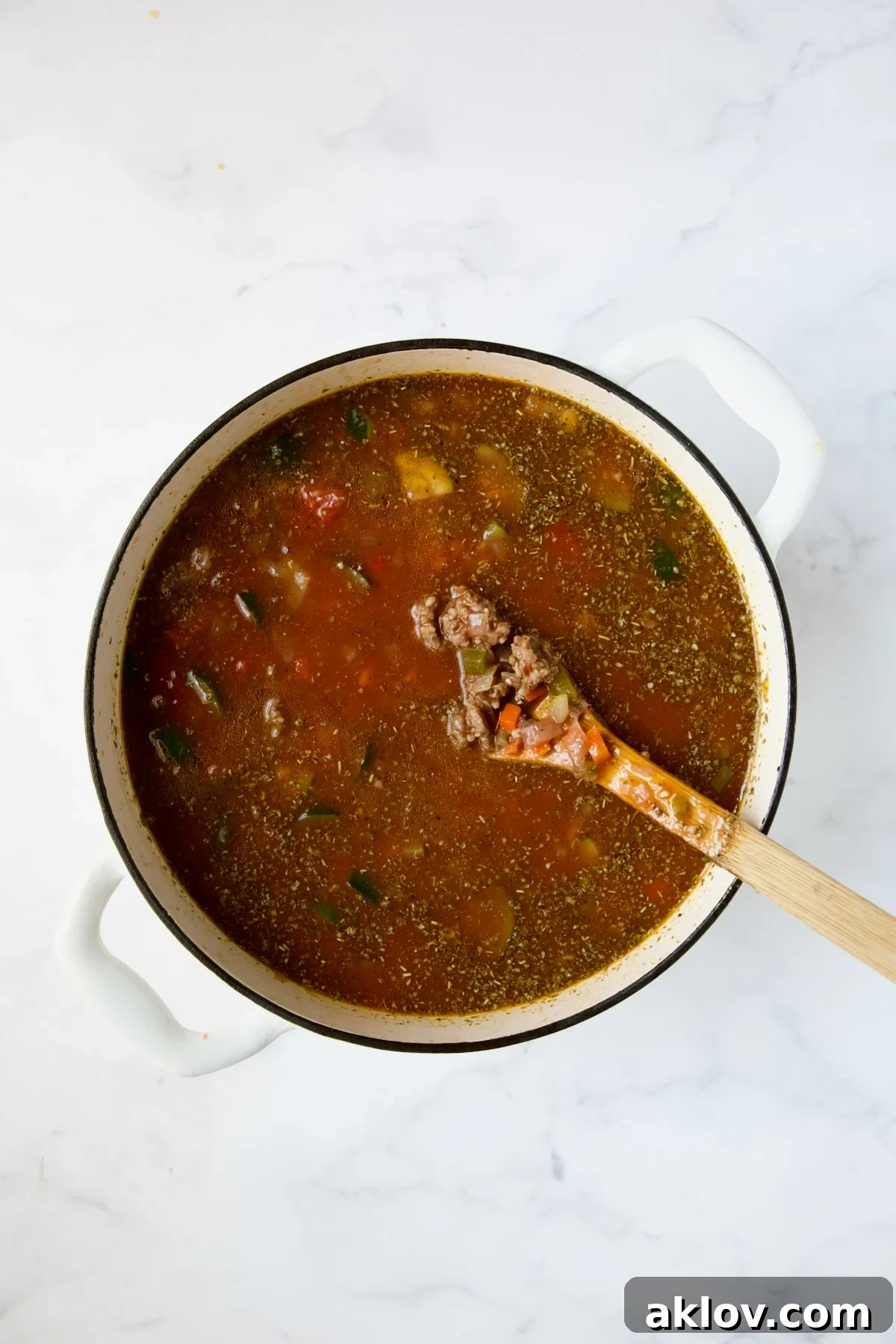 A wooden spoon stirring the simmering Italian sausage soup in a white Dutch oven.