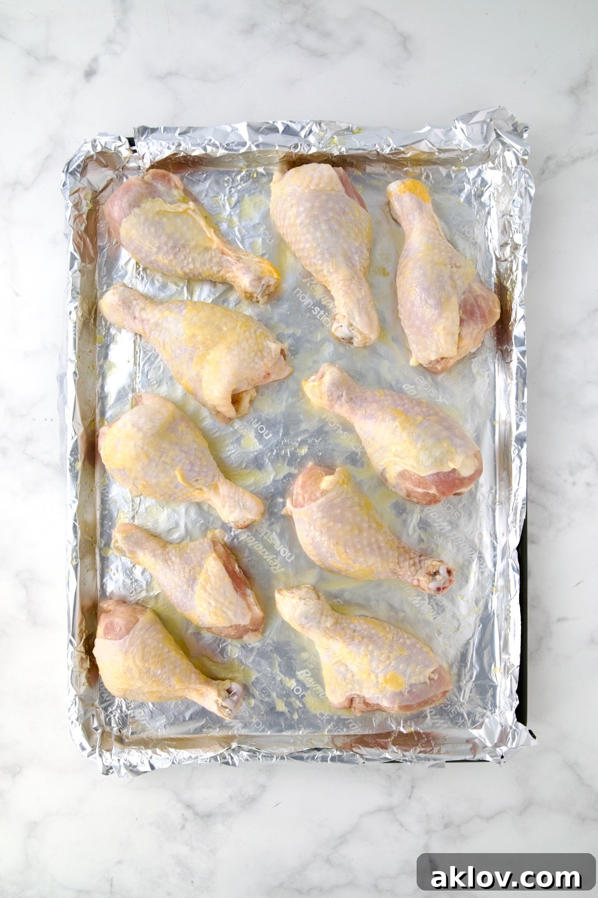 Ten uncooked, unseasoned chicken drumsticks arranged neatly on an aluminum foil-lined rimmed sheet pan.