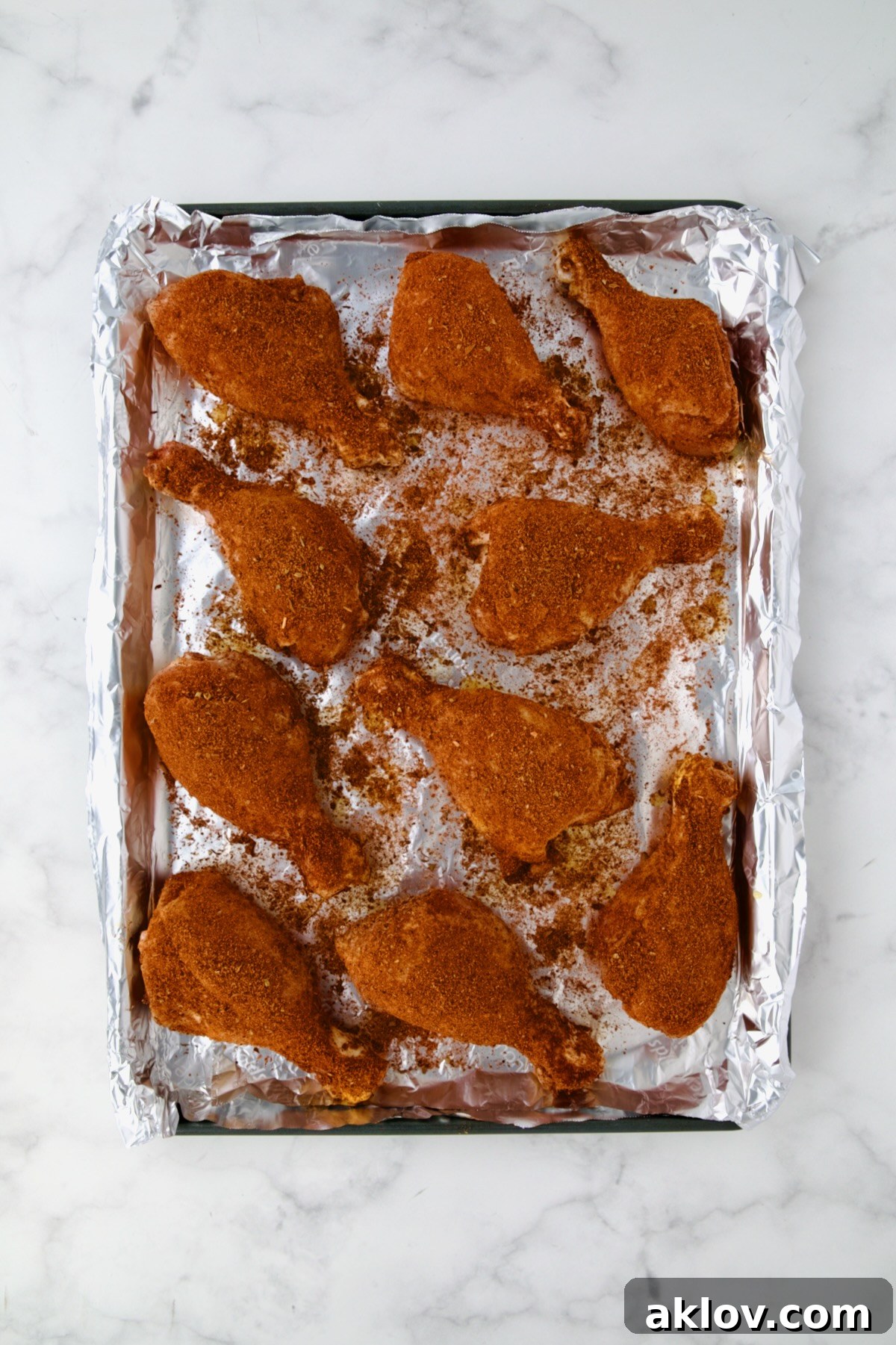 Ten uncooked, seasoned chicken drumsticks laid out on an aluminum foil-lined rimmed sheet pan, ready for baking.