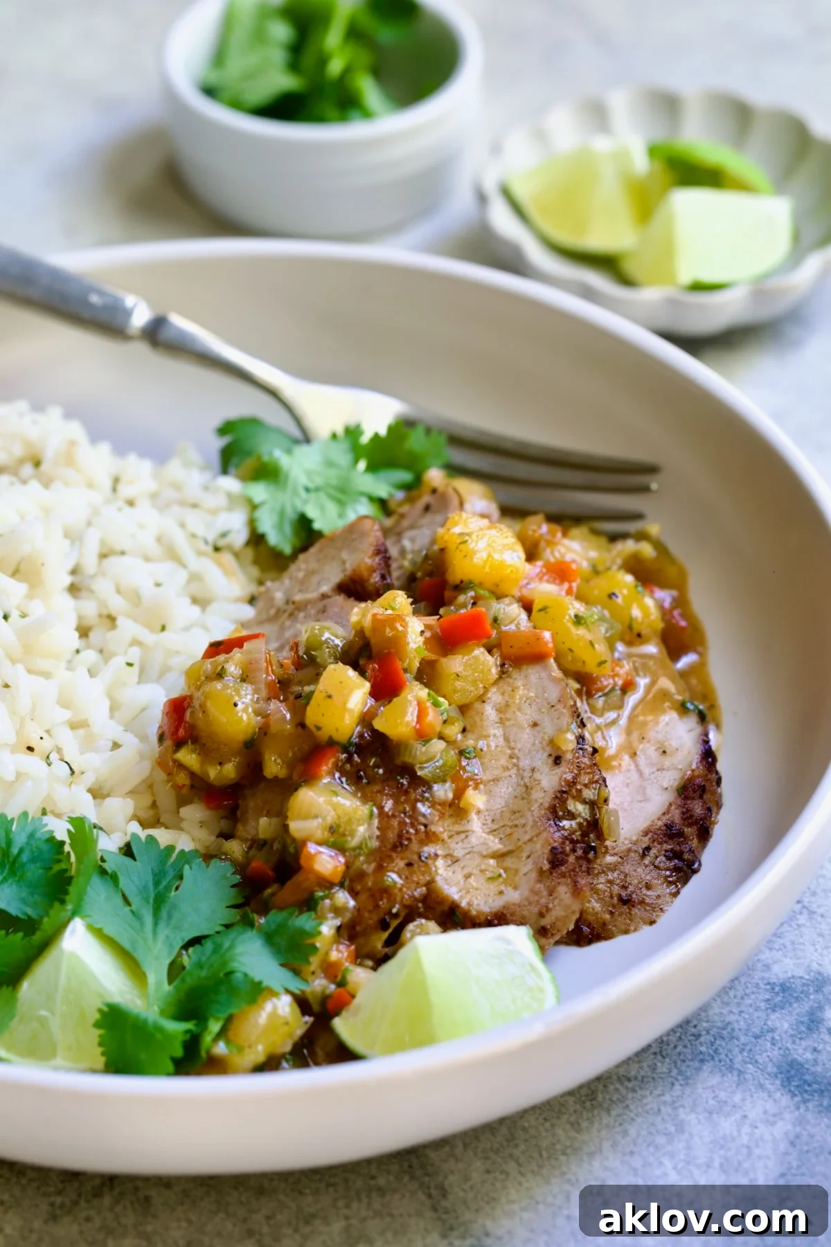 One serving of sliced and sauced Caribbean Pork Tenderloin in white dinner bowl garnished with cilantro and lime wedges.
