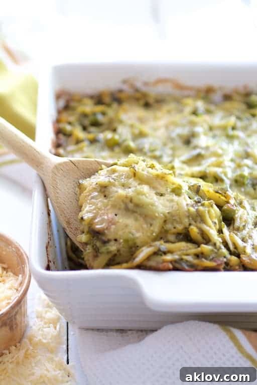 Photo of Baked Orzo with Pesto Peas Prosciutto and Mascarpone in white baking dish with some being scooped out with wooden spoon.