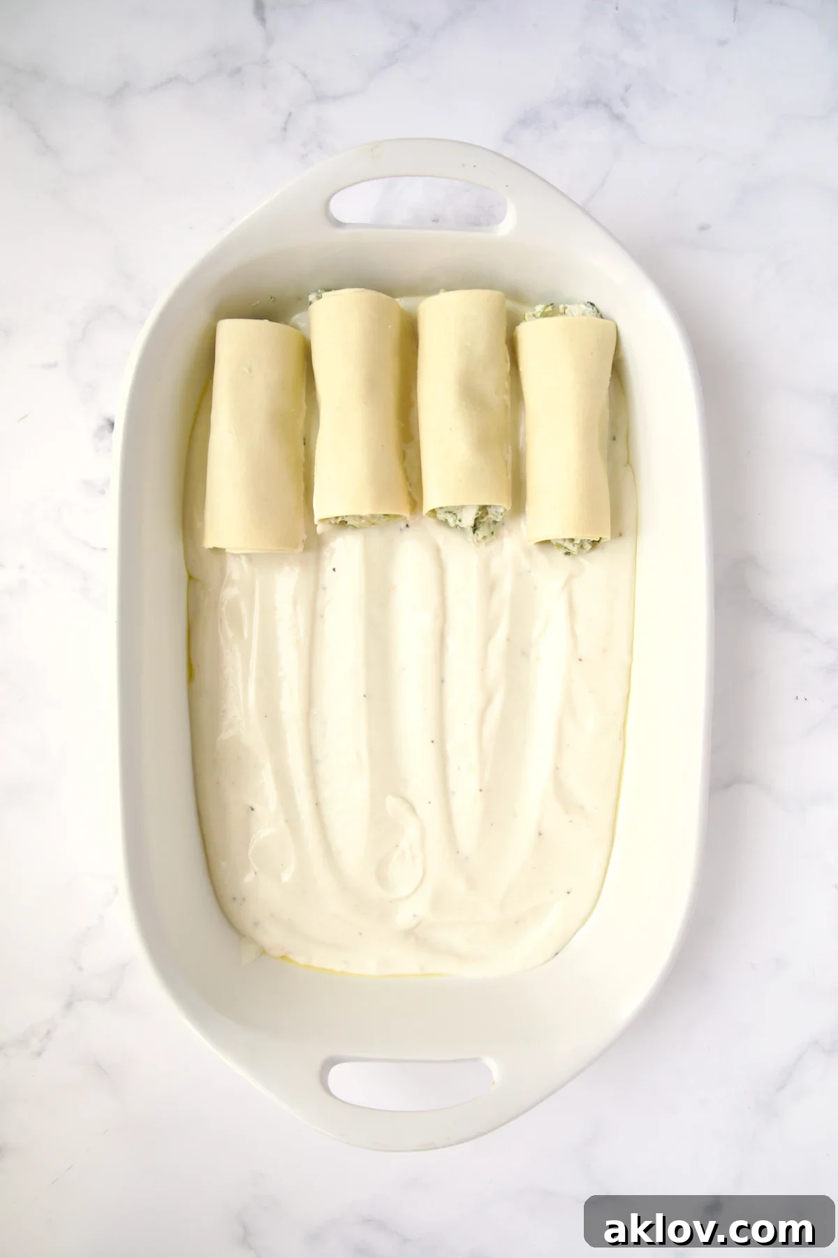 Four cannelloni set on bechamel sauce in white oval baking dish.