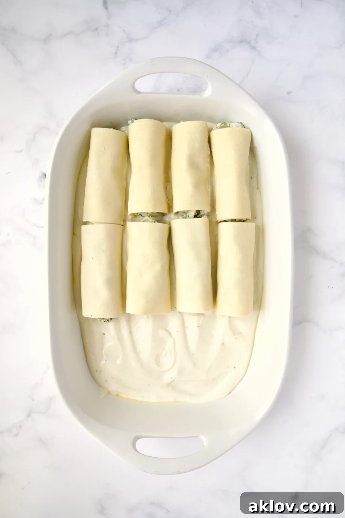 Eight cannelloni set on bechamel sauce in white oval baking dish.
