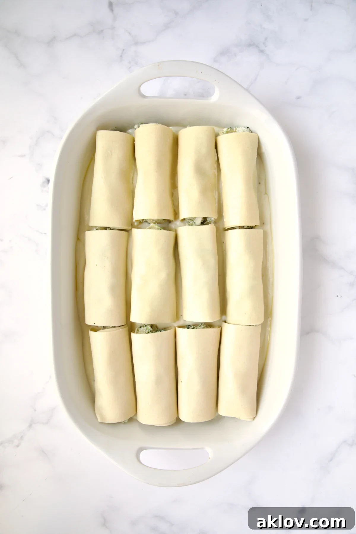 12 cannelloni set on bechamel sauce in white oval baking dish.