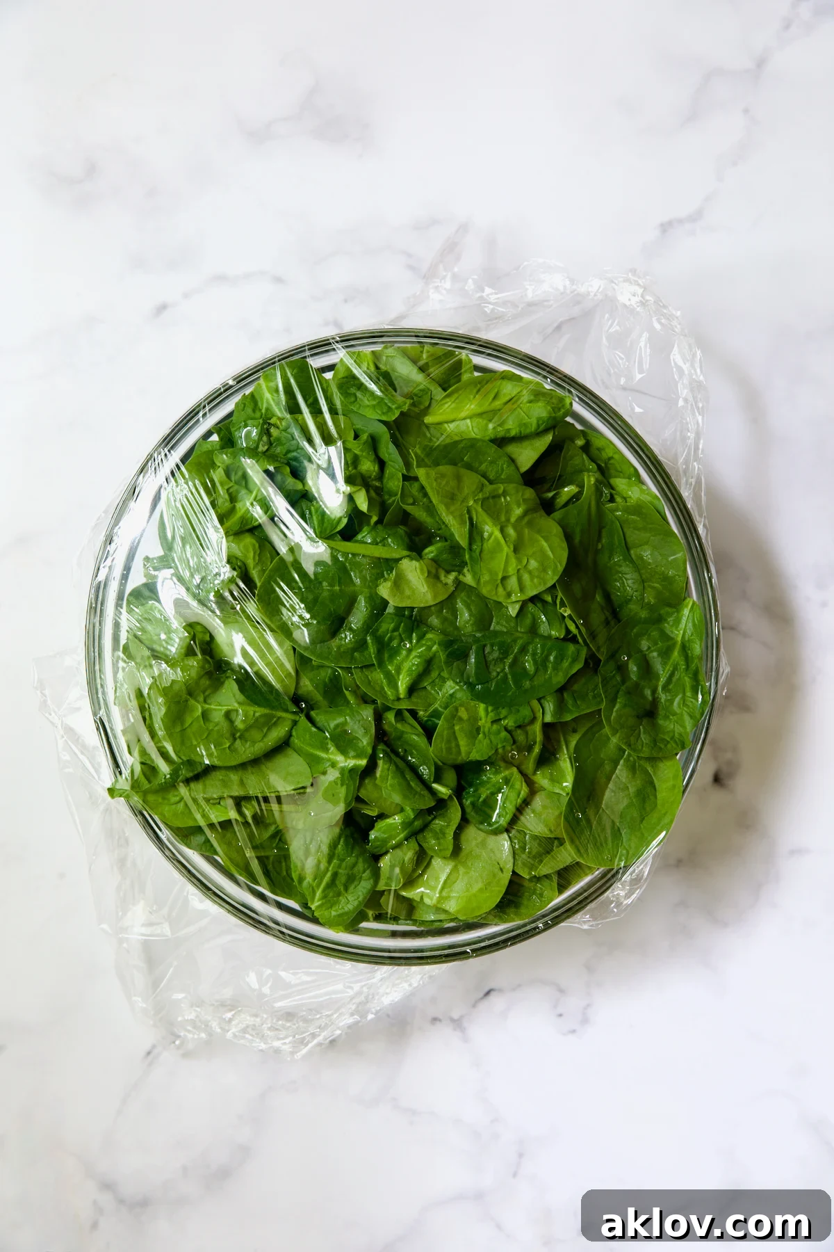 Glass bowl full of spinach with plastic wrap over the top.