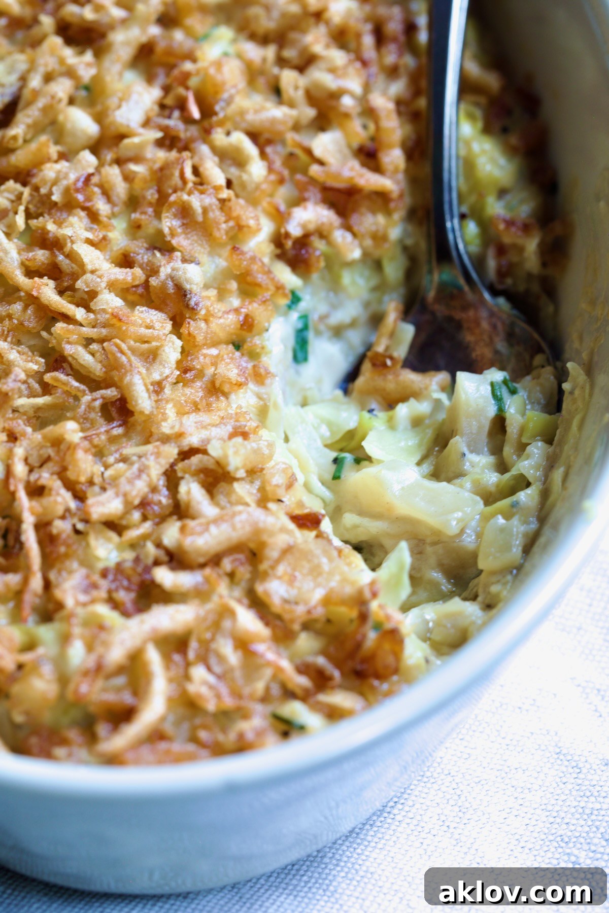 A close-up view of the Creamed Cabbage Casserole in a white oval baking dish, highlighting its creamy texture and the golden-brown French-fried onion topping, with an ornate serving spoon.