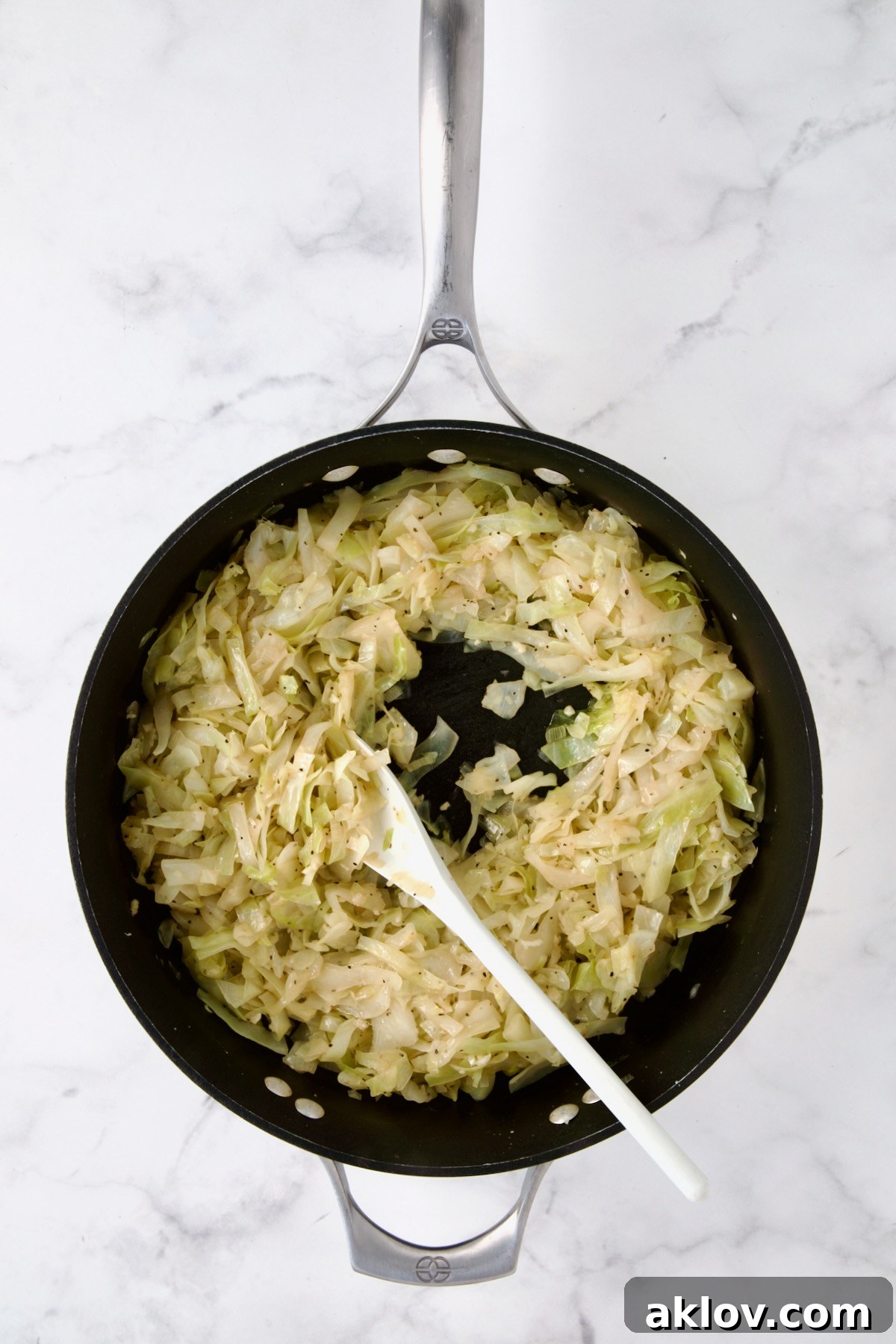 Tender, braised cabbage being stirred in a nonstick skillet with a white mixing spoon, coated in a rich, buttery broth.