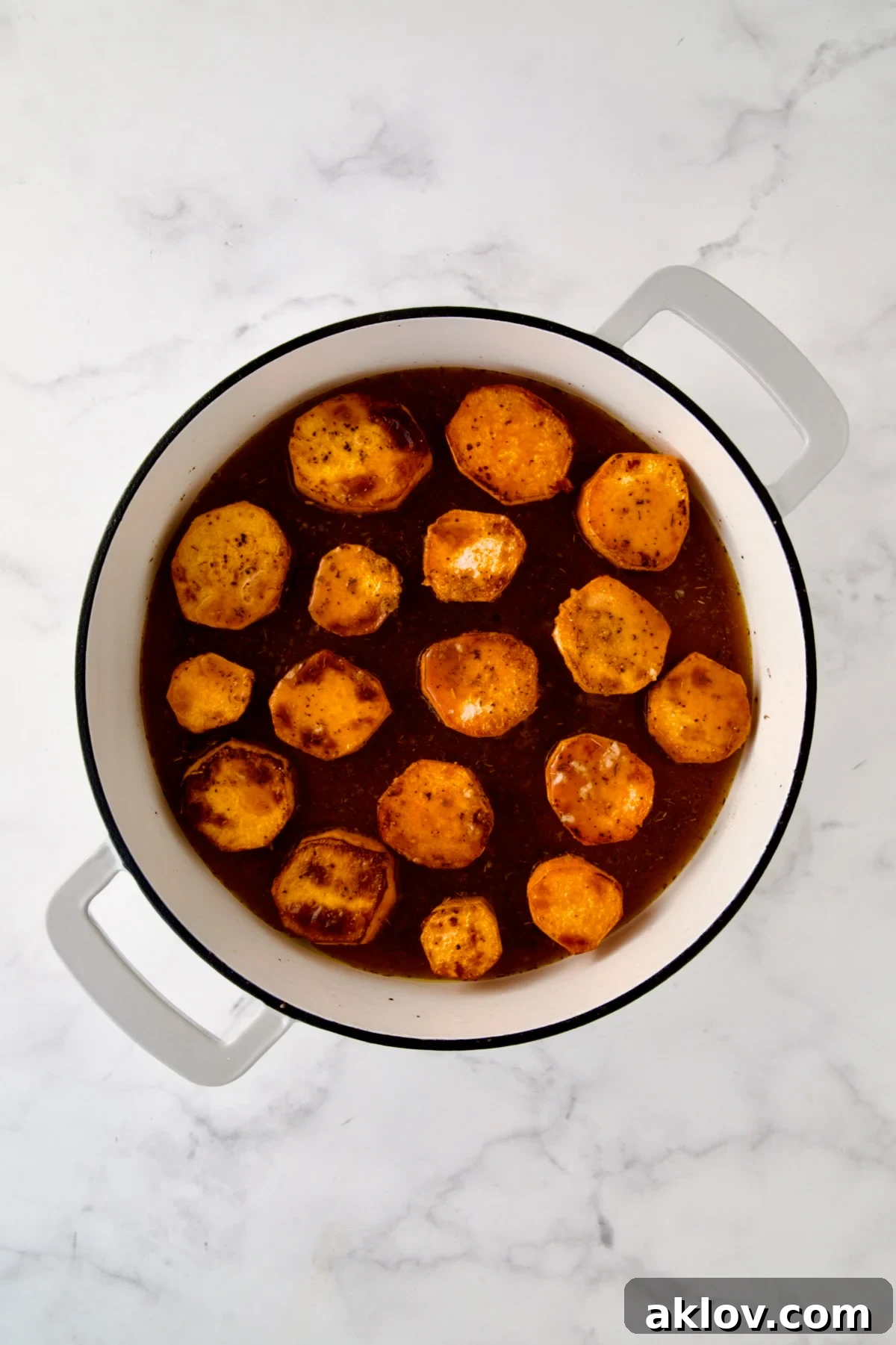 Rounds of browned sweet potatoes in white enamel cast iron skillet in herb and broth blend.