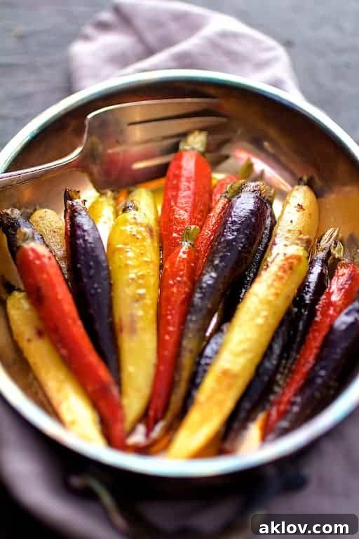 A close-up of Curry Roasted Carrots with Honey Browned Butter in an elegant serving dish, with a silver serving fork resting on a gray linen napkin, highlighting the textures and colors.