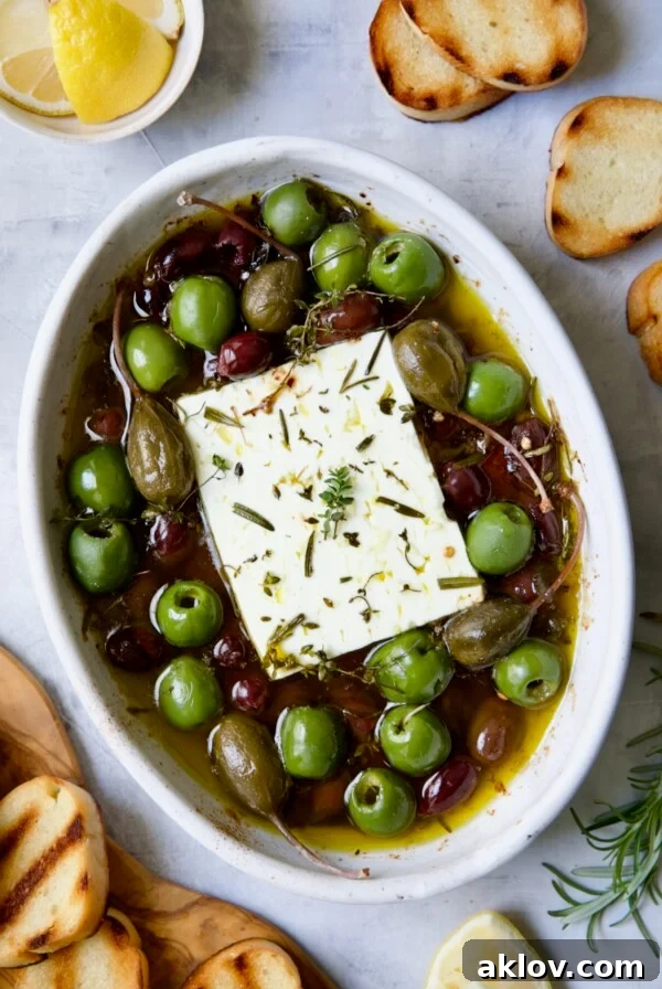 Baked Feta Cheese and Olives in white oval baking dish with serving knife surrounded by grilled bread slices.