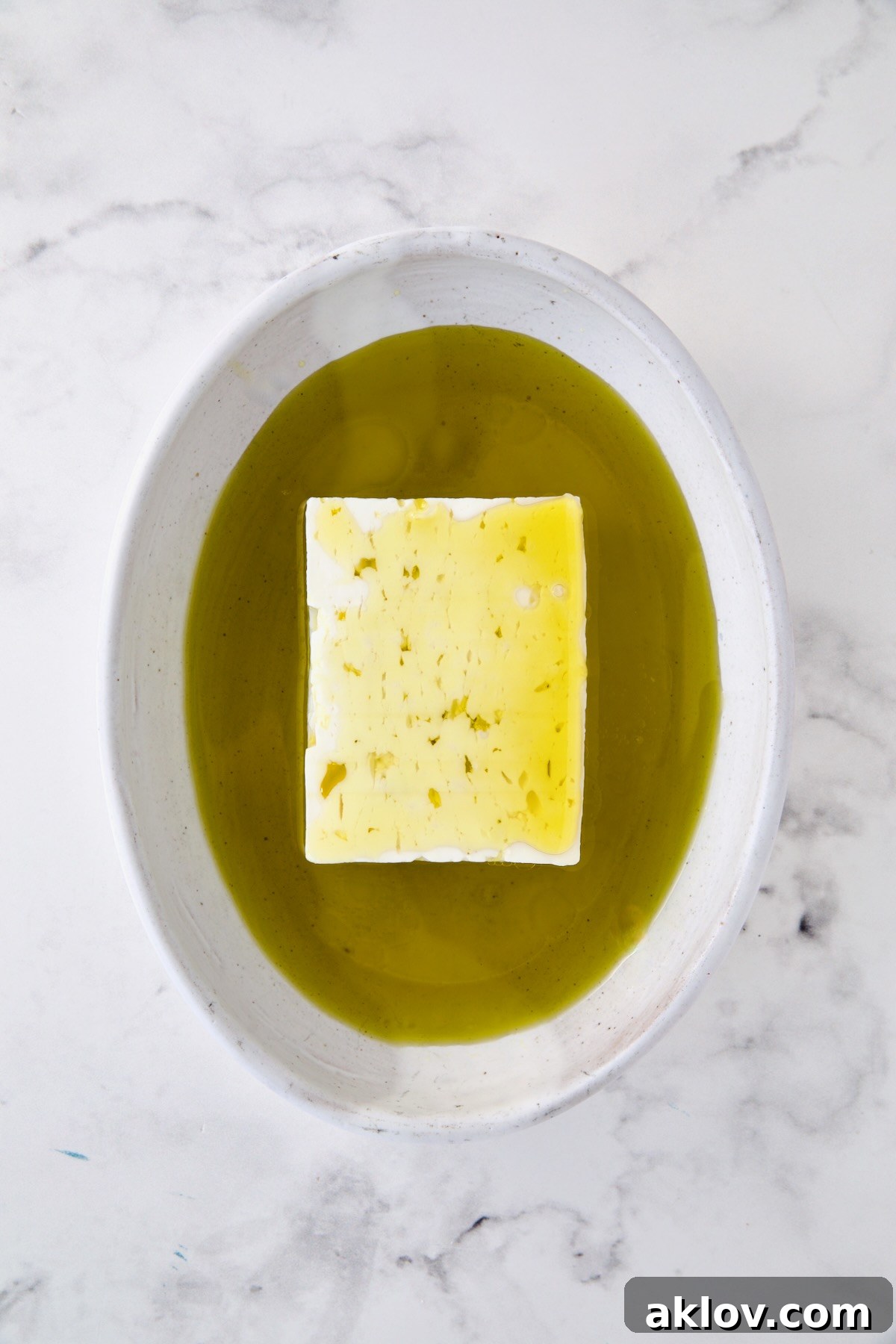 Block of feta cheese in oval white baking dish with olive oil poured over it.