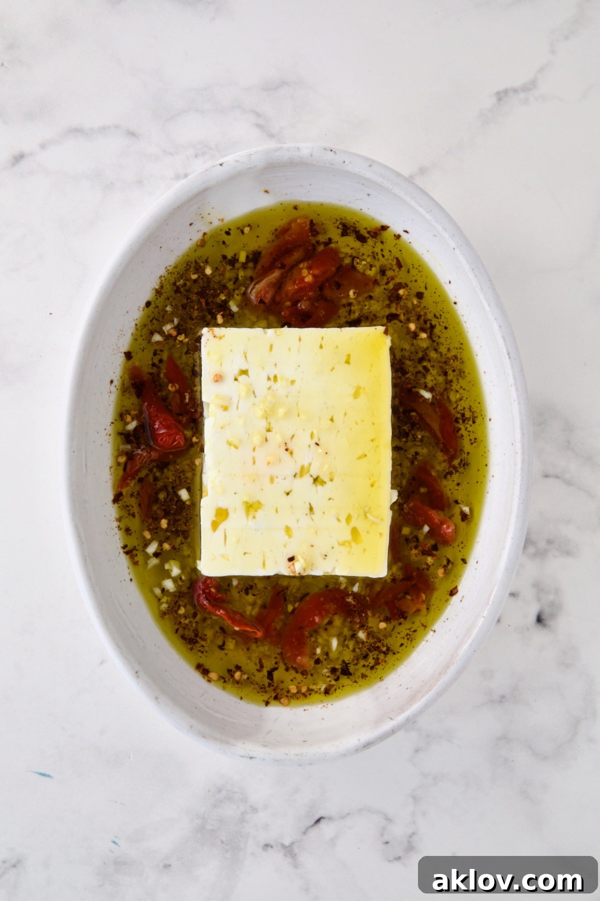 Block of feta cheese in oval white baking dish with olive oil poured over it and garlic, crushed red pepper flakes and sun-dried tomatoes added.