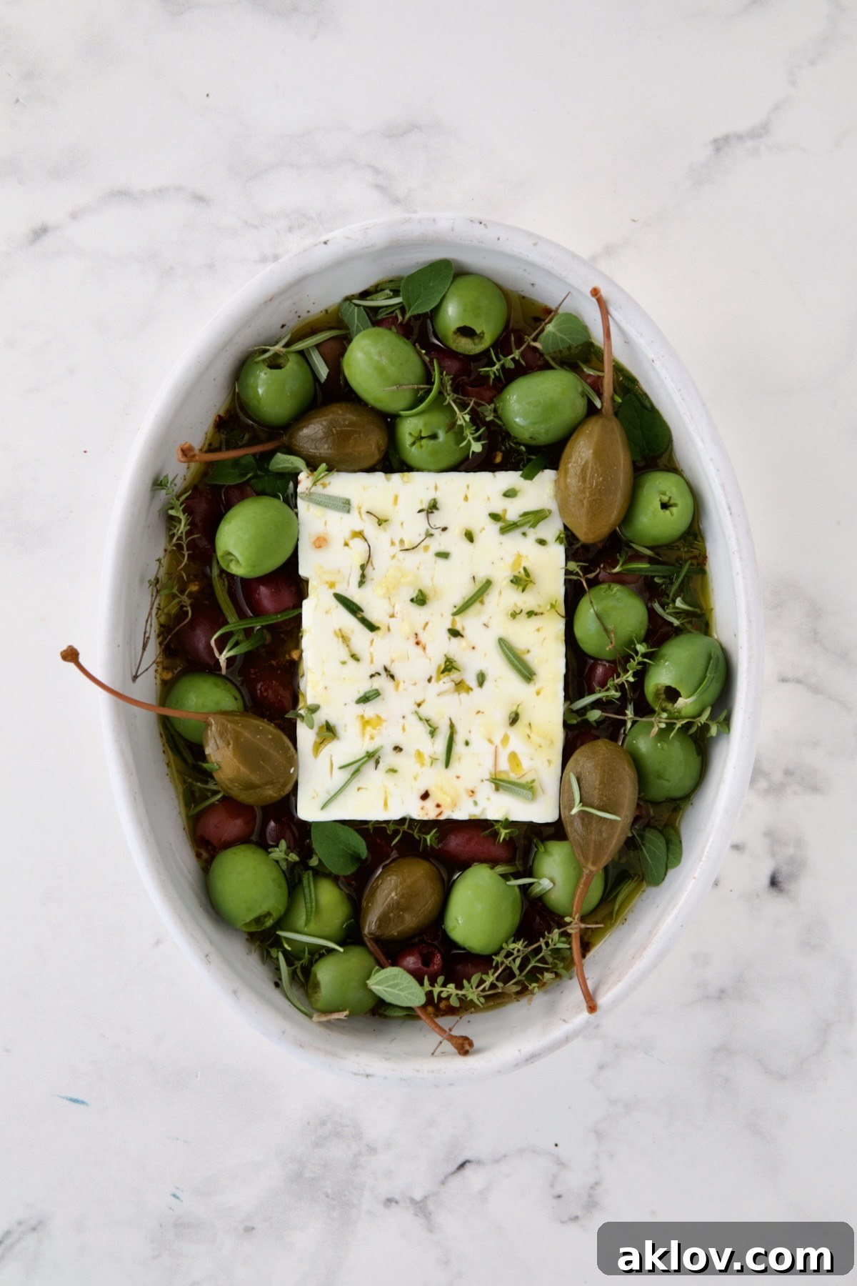 Block of feta cheese in oval white baking dish with olive oil poured over it and garlic, crushed red pepper flakes, sun-dried tomatoes, olives and caper berries added.