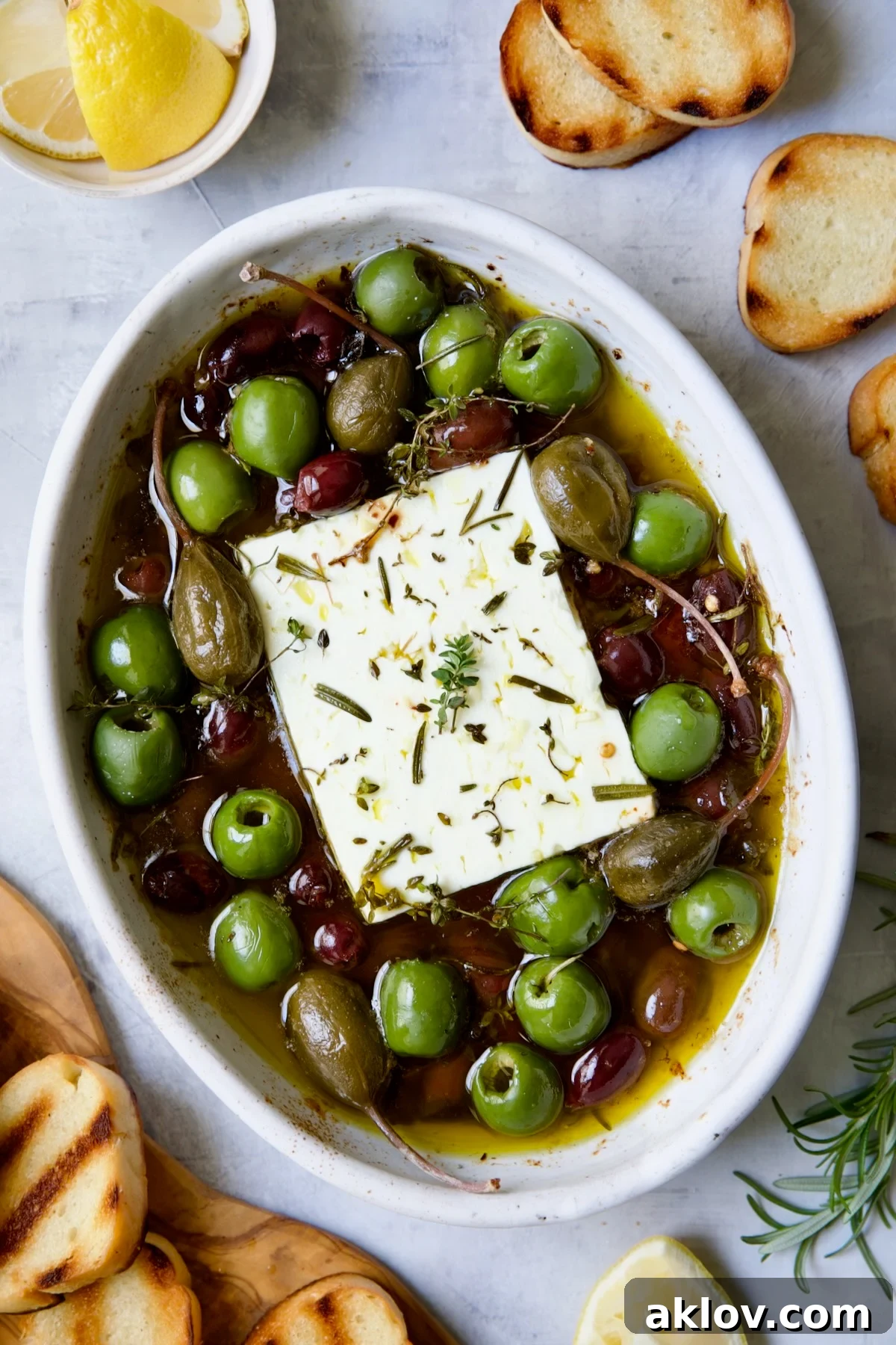 Baked Feta Cheese and Olives in white oval baking dish with serving knife surrounded by grilled bread slices.