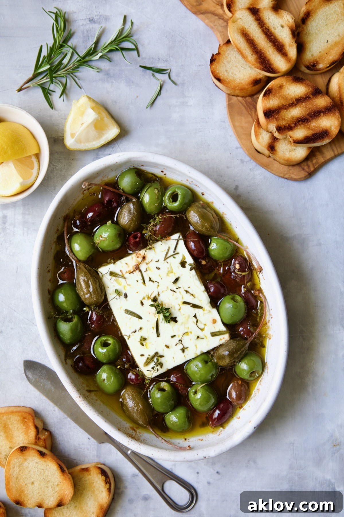 Baked Feta Cheese and Olives in white oval baking dish with serving knife surrounded by grilled bread slices and lemon wedges.