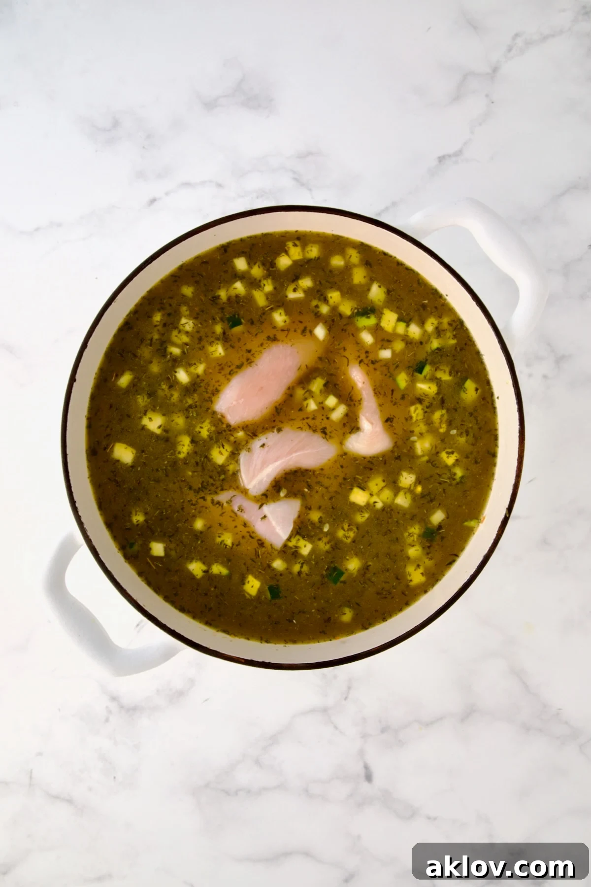 Raw chicken added to broth and vegetables in white Dutch oven.