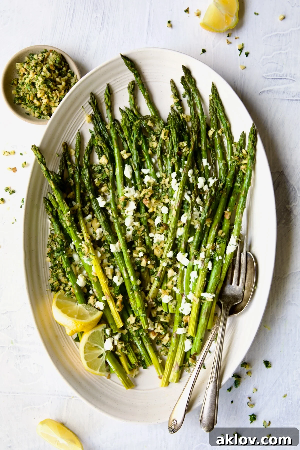 Roasted Asparagus with Walnut Lemon Gremolata and Goat Cheese artfully arranged on an oval white platter, garnished with fresh lemon wedges and serving utensils.